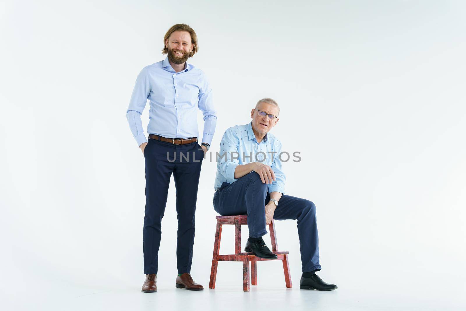 Father sits on the chair and son stands on white background, both men look into the camera, an elderly man in glasses with diopters, a stylish young man with a beard and long hair. High quality photo