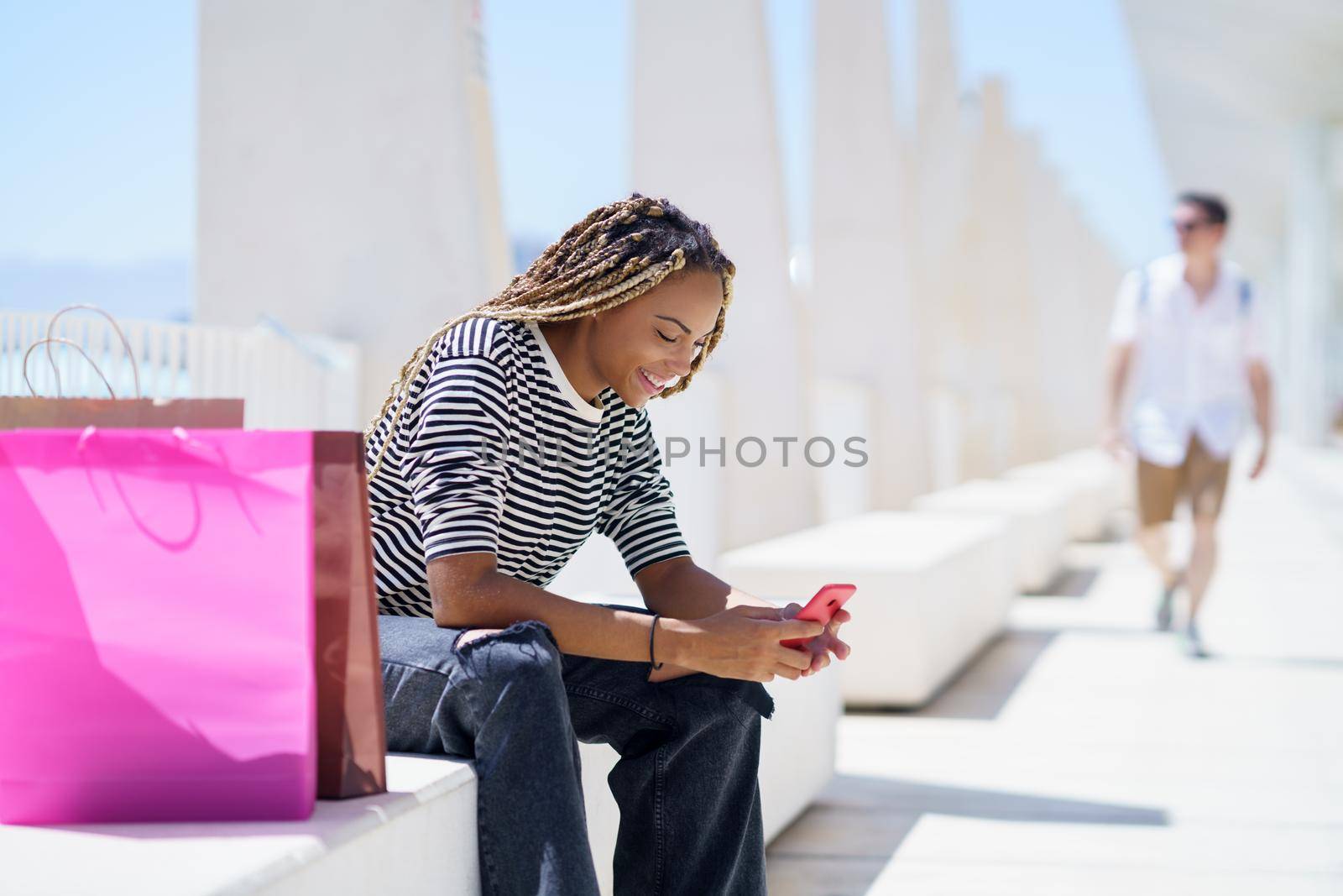 Young black woman using her smartphone sitting on a bench in the street.