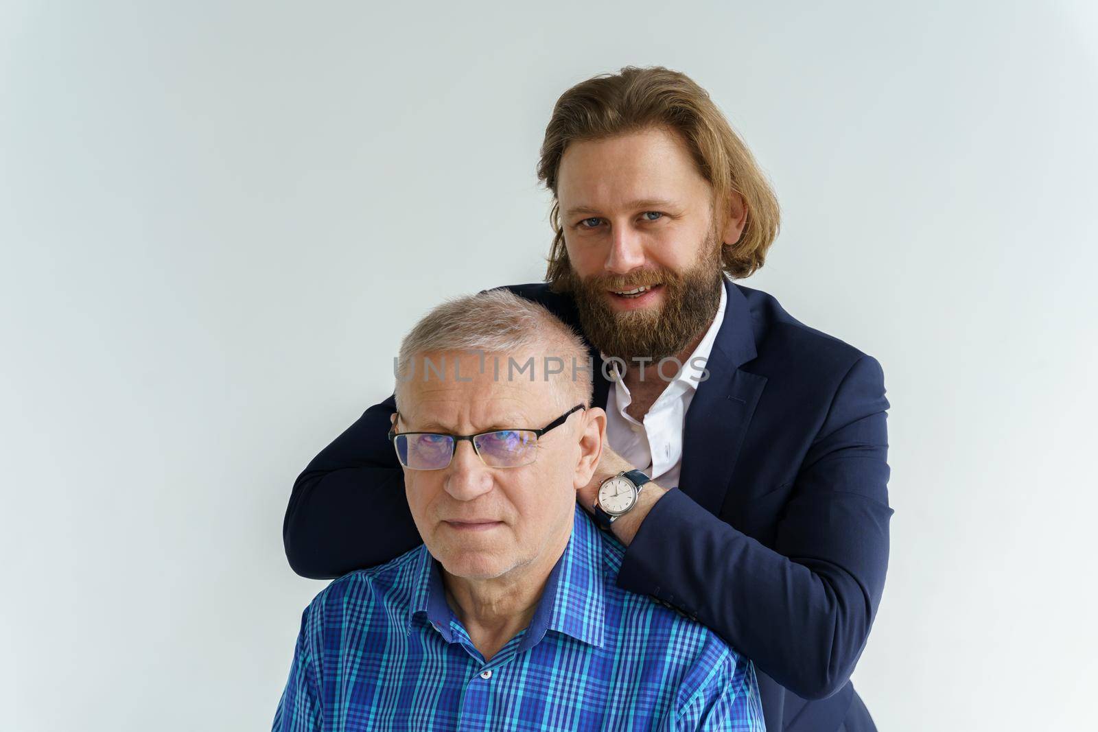 the son in a stylish suit leans on the shoulders of his father, Father and son in white background, both men look into the camera, an elderly man in glasses, young man with a beard and long hair by vladimirdrozdin