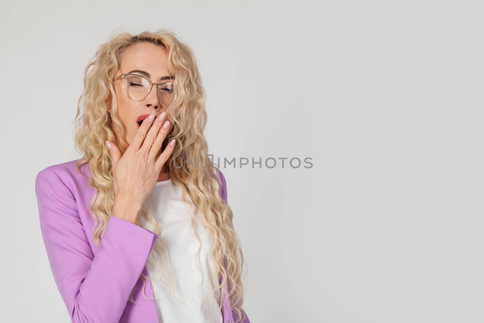 Snapshot of a sleepy attractive Caucasian woman blonde with curly hair wearing glasses, feeling tired after a night without sleep, yawning, covering her open mouth with her palm