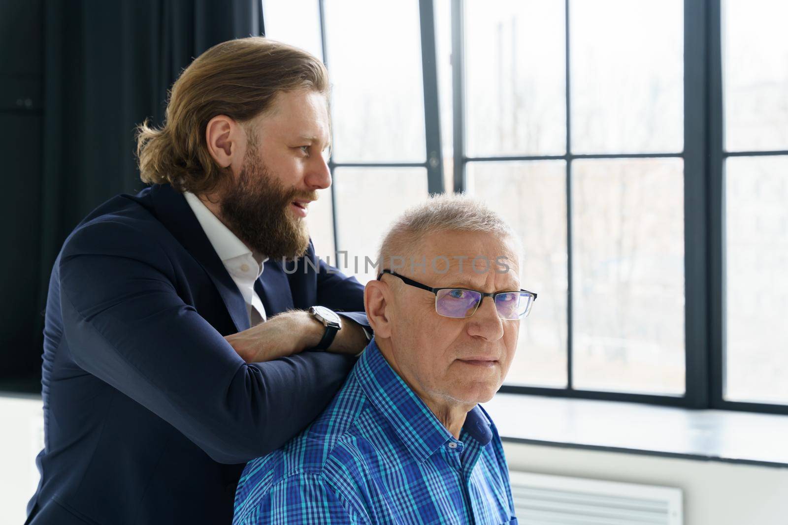 the son in a stylish suit leans on the shoulders of his father, Father and son in huge window background, both men look into the camera, an elderly man in glasses, young man with a beard and long hair. High quality photo