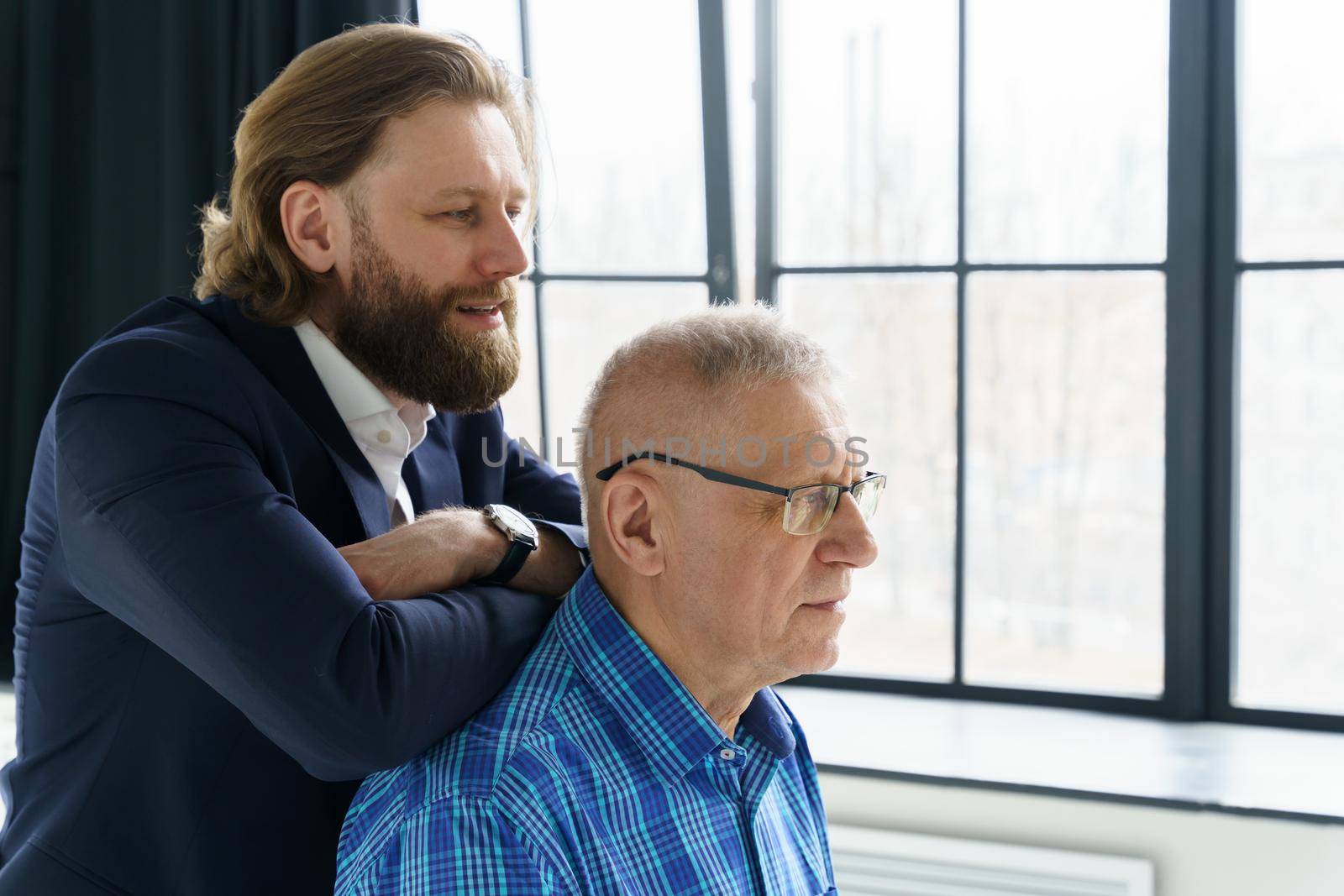 the son in a stylish suit leans on the shoulders of his father, Father and son in huge window background, both men look into the camera, an elderly man in glasses, young man with a beard and long hair. High quality photo