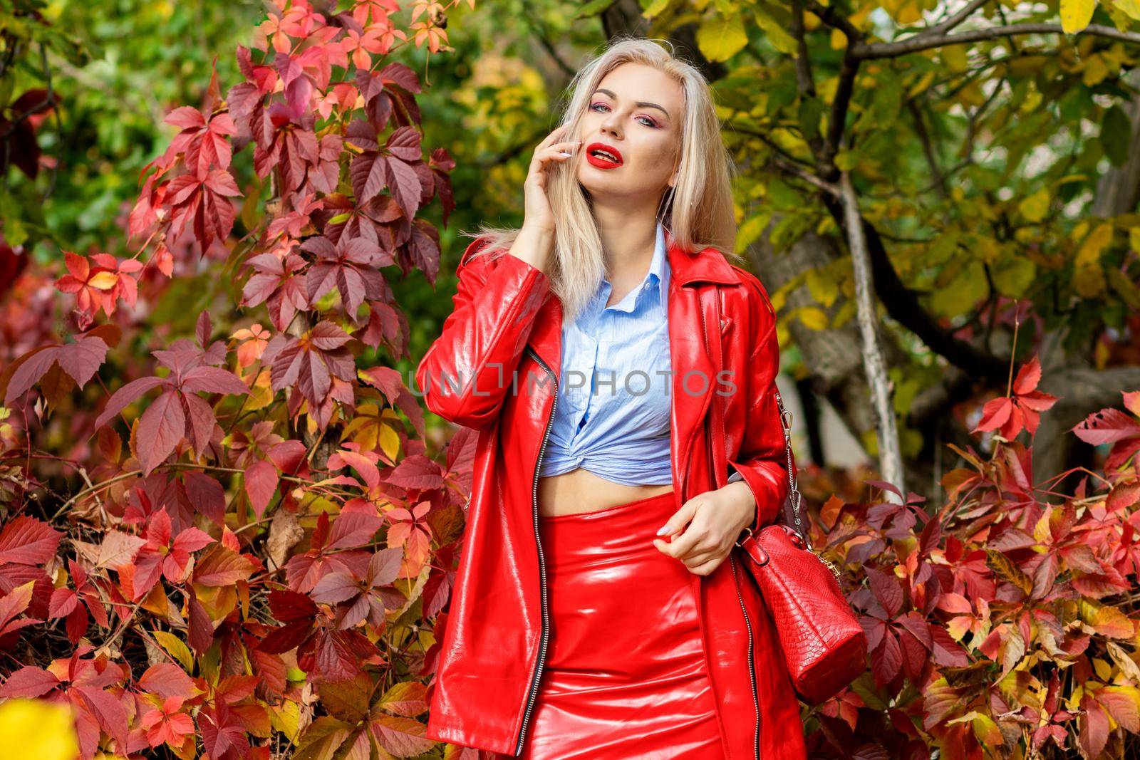 A fashionable woman wears a trendy white and blue shirt and red skirt and leather jacket on a city street. Stylish young blonde lady with bright makeup.