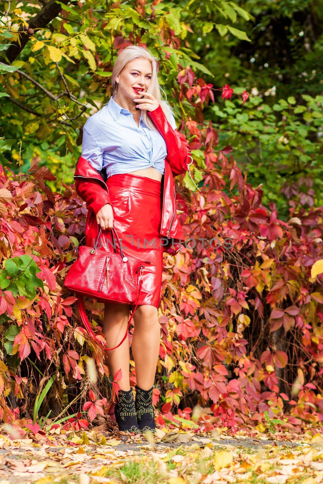 A fashionable woman wears a trendy white and blue shirt and red skirt and leather jacket on a city street. Stylish young blonde lady with bright makeup.