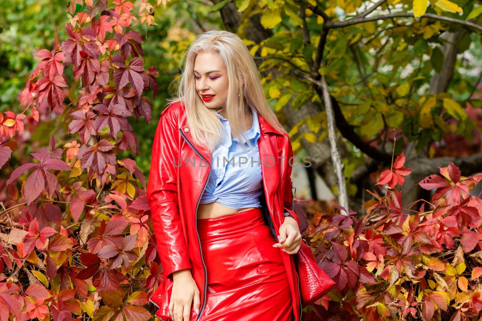 A fashionable woman wears a trendy white and blue shirt and red skirt and leather jacket on a city street. Stylish young blonde lady with bright makeup.