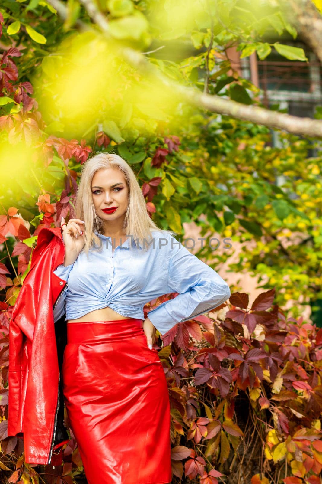 A fashionable woman wears a trendy white and blue shirt and red skirt and leather jacket on a city street. Stylish young blonde lady with bright makeup.