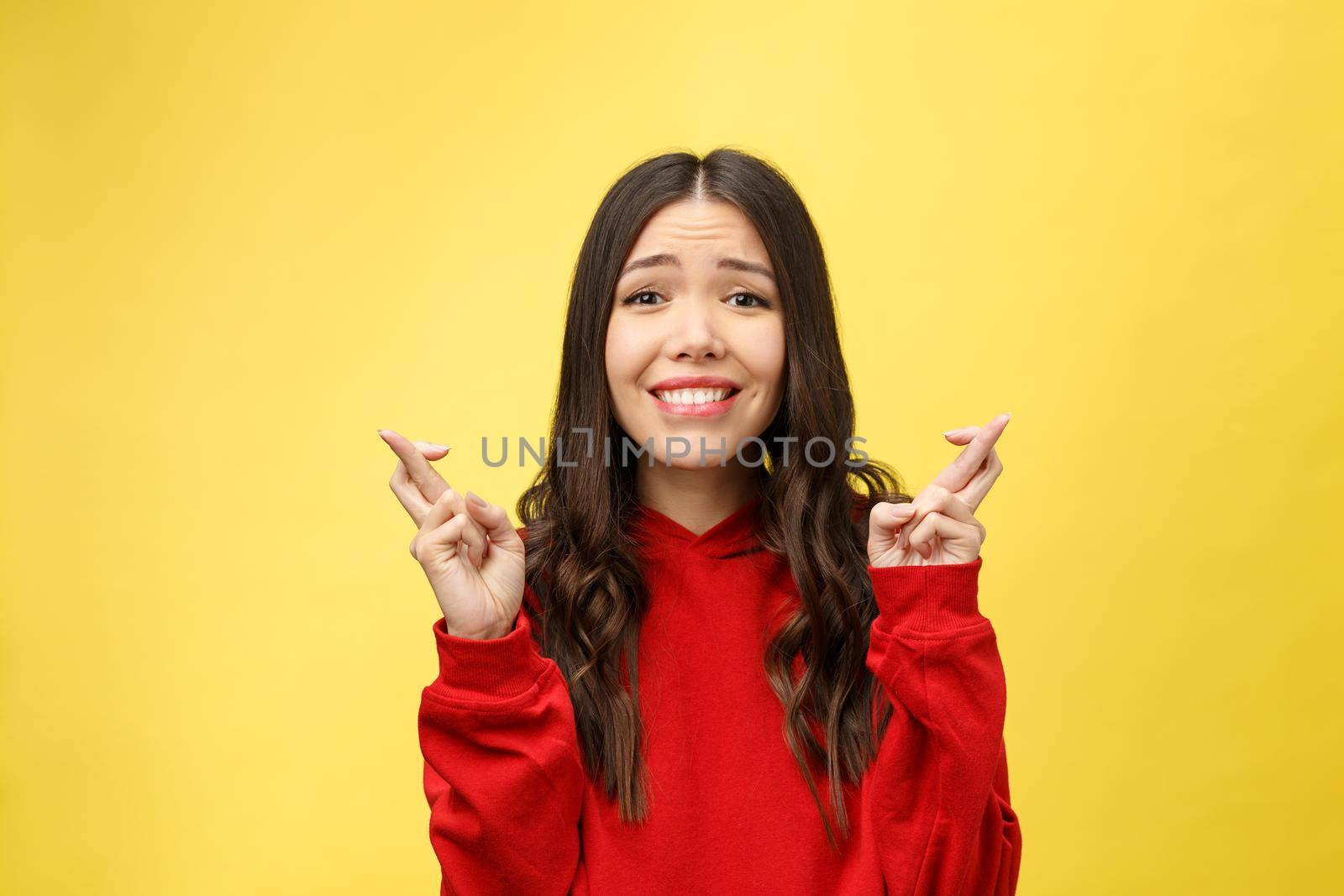 Intrigued woman in praying with crossed fingers and looking away over yellow background.