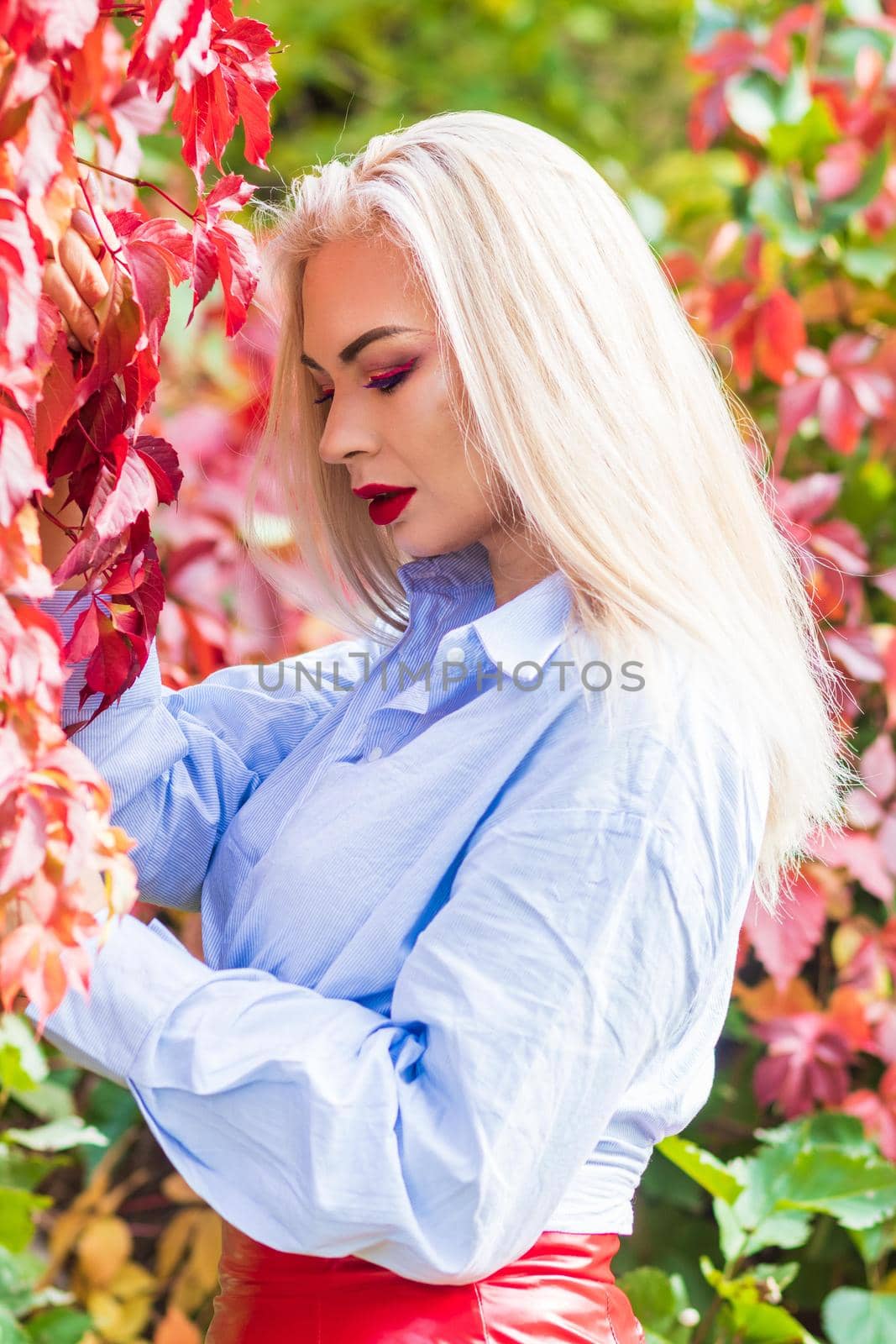 A fashionable woman wears a trendy white and blue shirt and red skirt and leather jacket on a city street. Stylish young blonde lady with bright makeup.