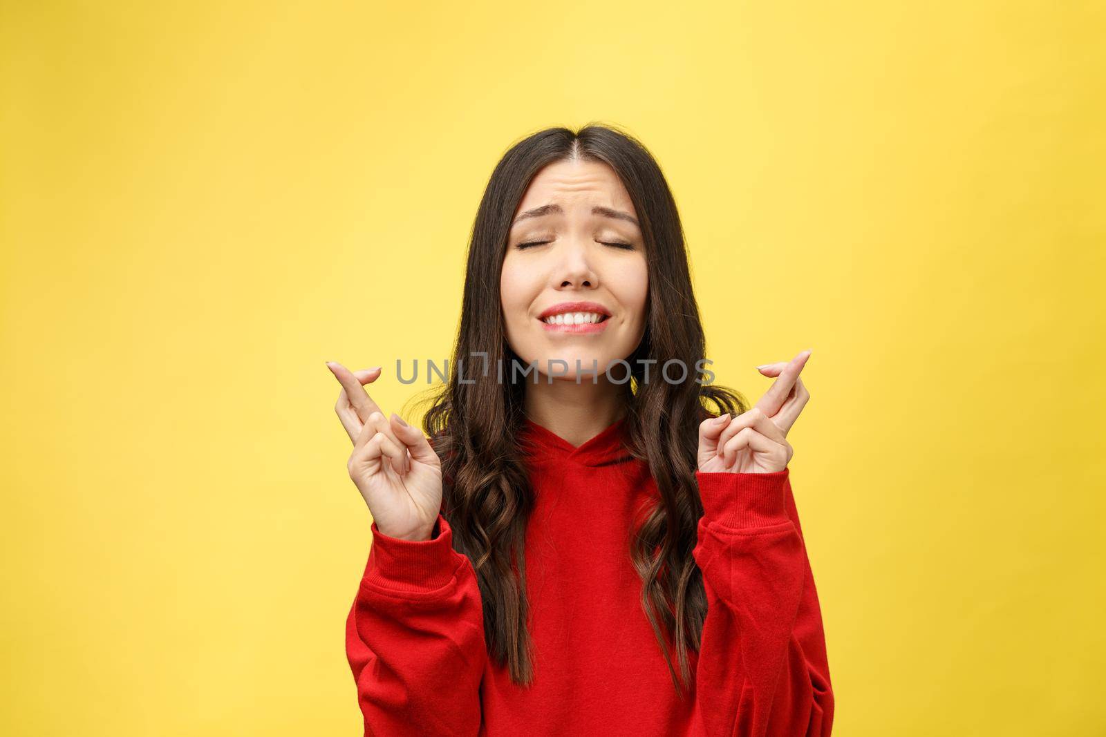 Intrigued woman in praying with crossed fingers and looking away over yellow background.