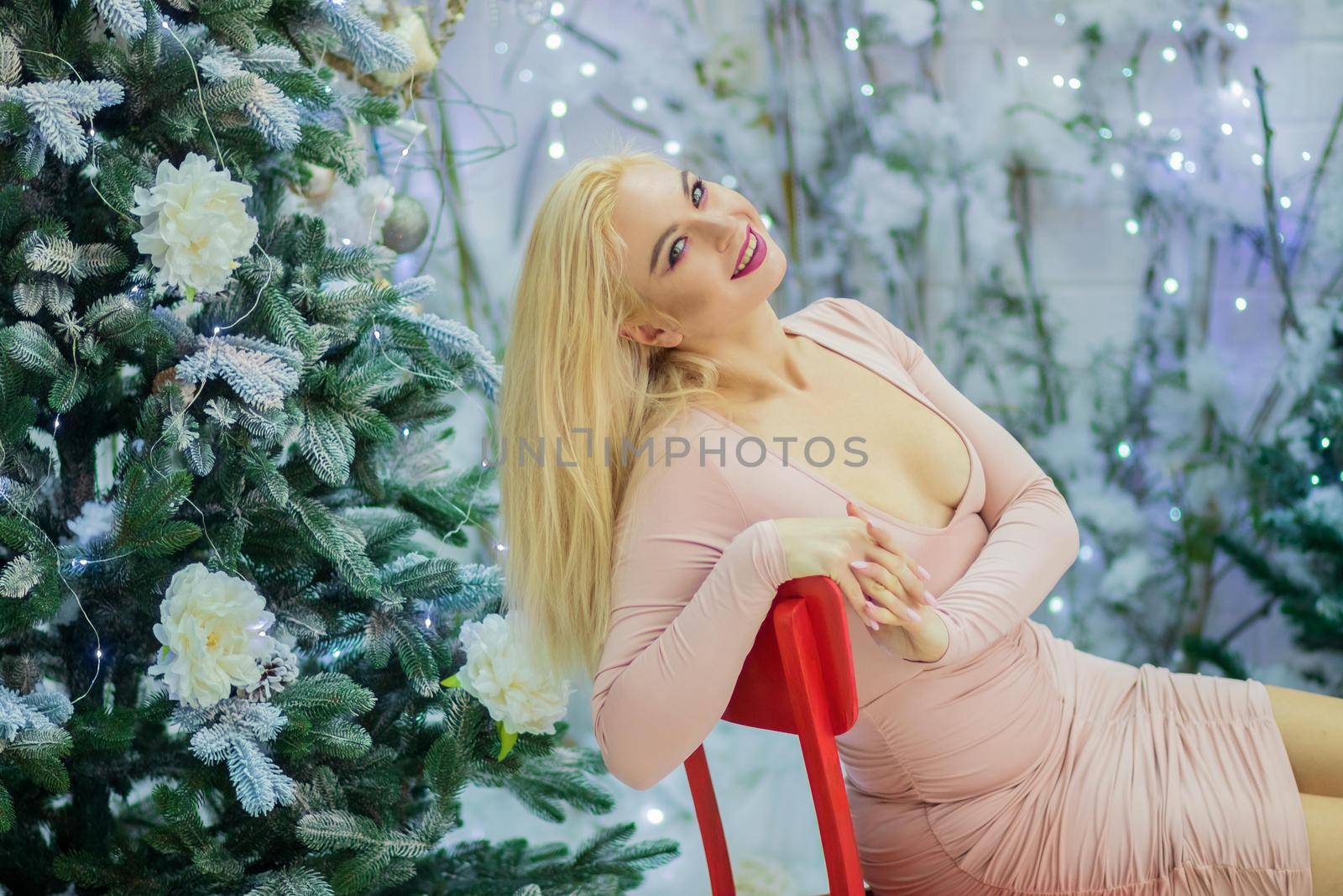 Charming blonde woman in beige sexy dress sitting on red chair, over background with Christmas tree. Christmas and New Year photo.