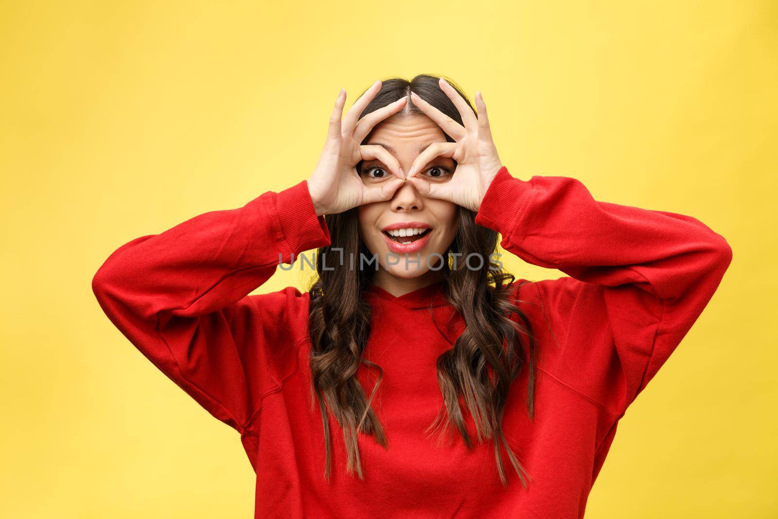 Close up portrait of beautiful joyful Caucasian female smiling, demonstrating white teeth, looking at the camera through fingers in okay gesture. Face expressions, emotions, and body language.