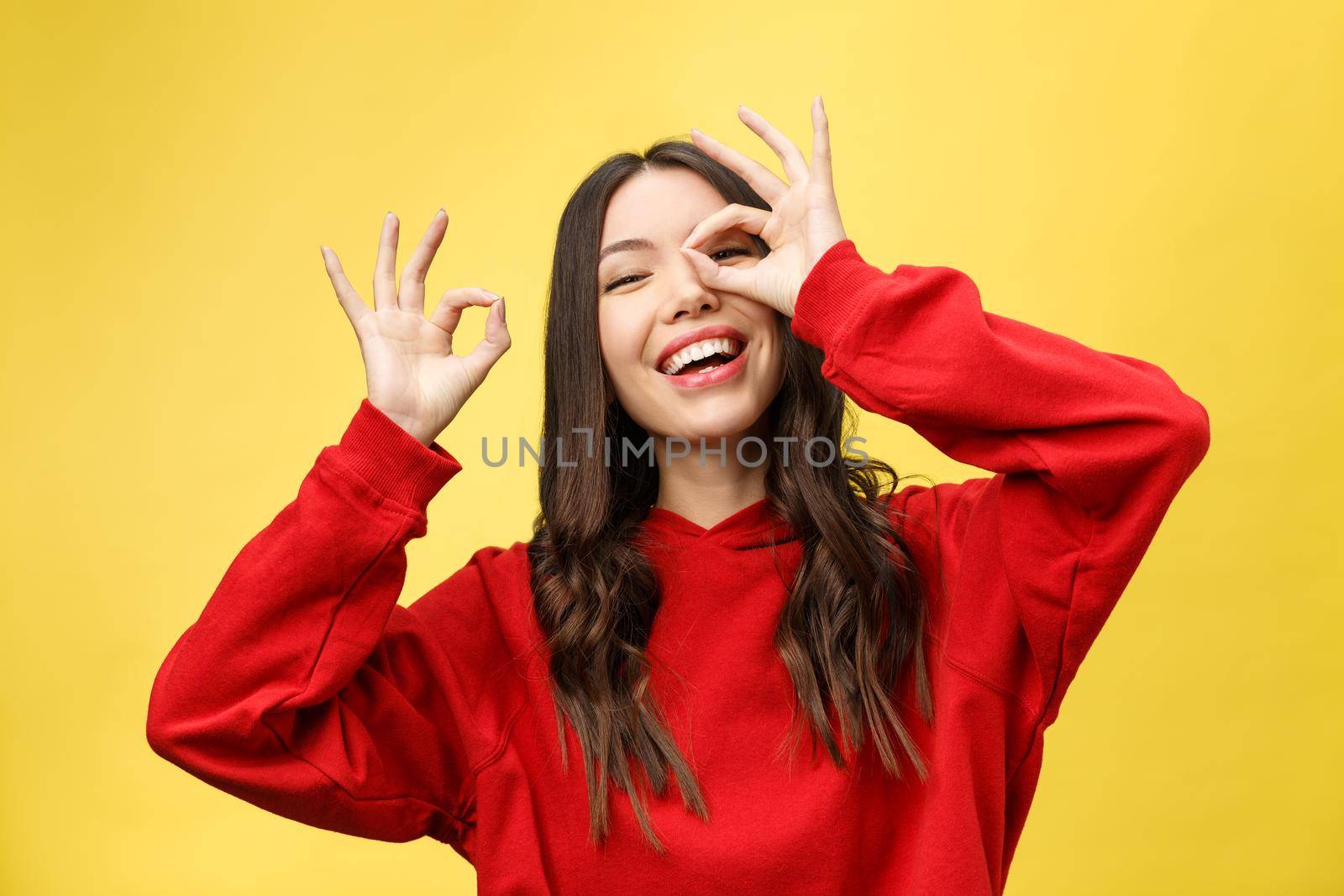 Close up portrait of beautiful joyful Caucasian female smiling, demonstrating white teeth, looking at the camera through fingers in okay gesture. Face expressions, emotions, and body language by Benzoix