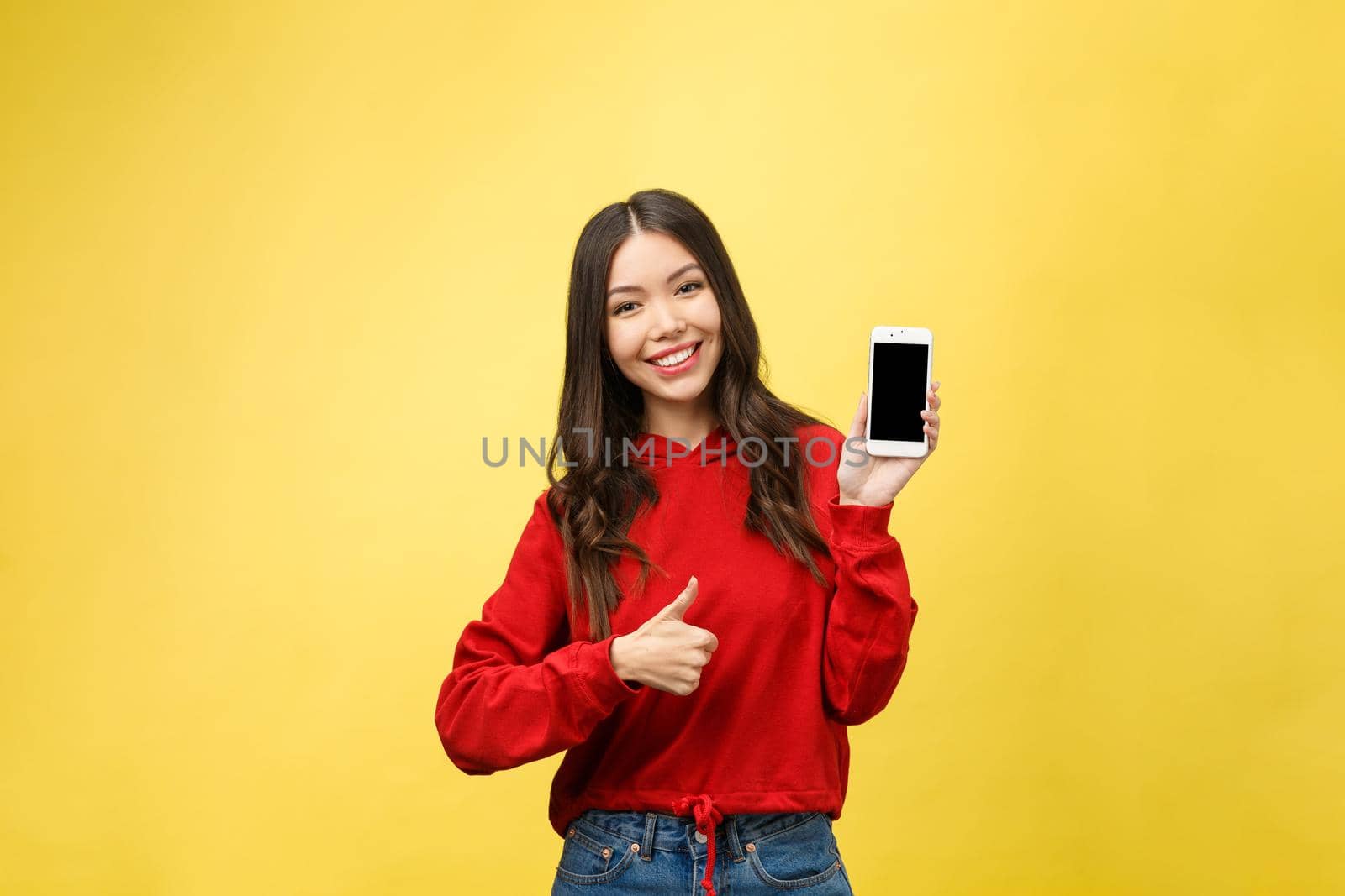 Portrait of a cheerful casual girl holding mobile phone and pointing finger away isolated over yellow background.