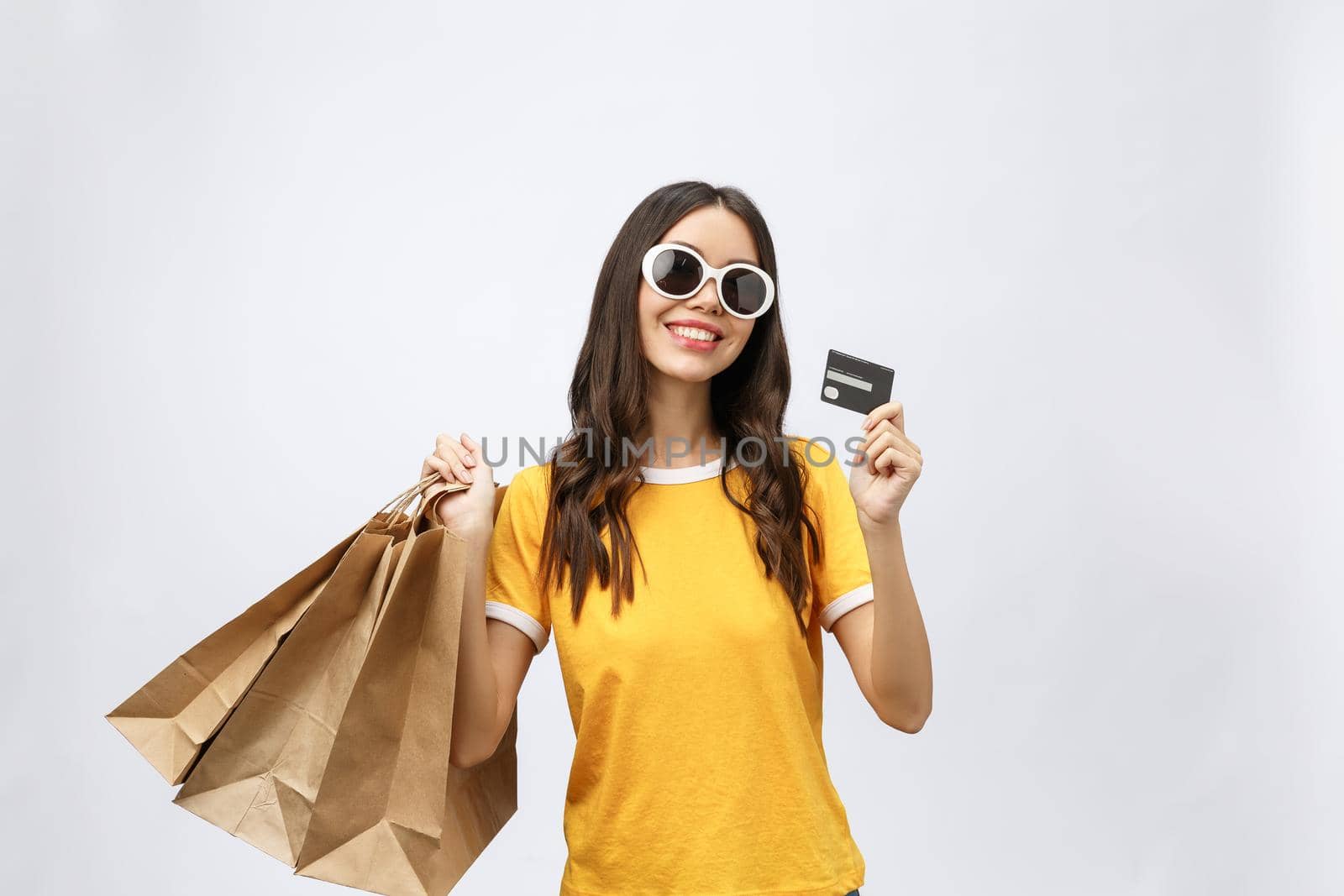 Close-up portrait of happy young brunette woman in sunglasses holding credit card and colorful shopping bags, looking at camera, isolated on white background by Benzoix