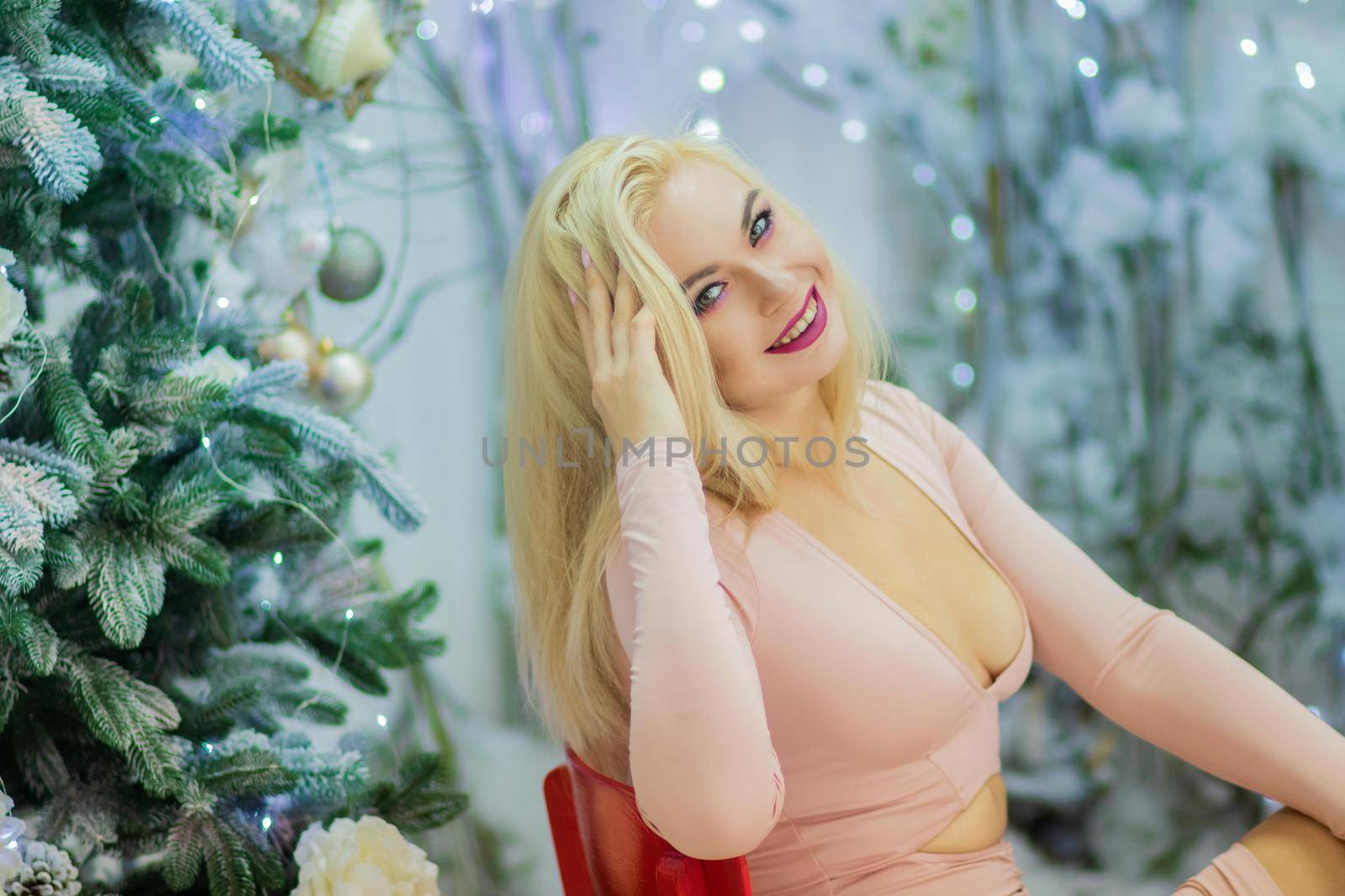Charming blonde woman in beige sexy dress sitting on red chair, over background with Christmas tree. Christmas and New Year photo.
