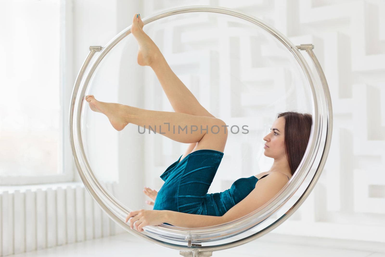 Young woman wearing blue dress lying in round glass chair in white room. Side view