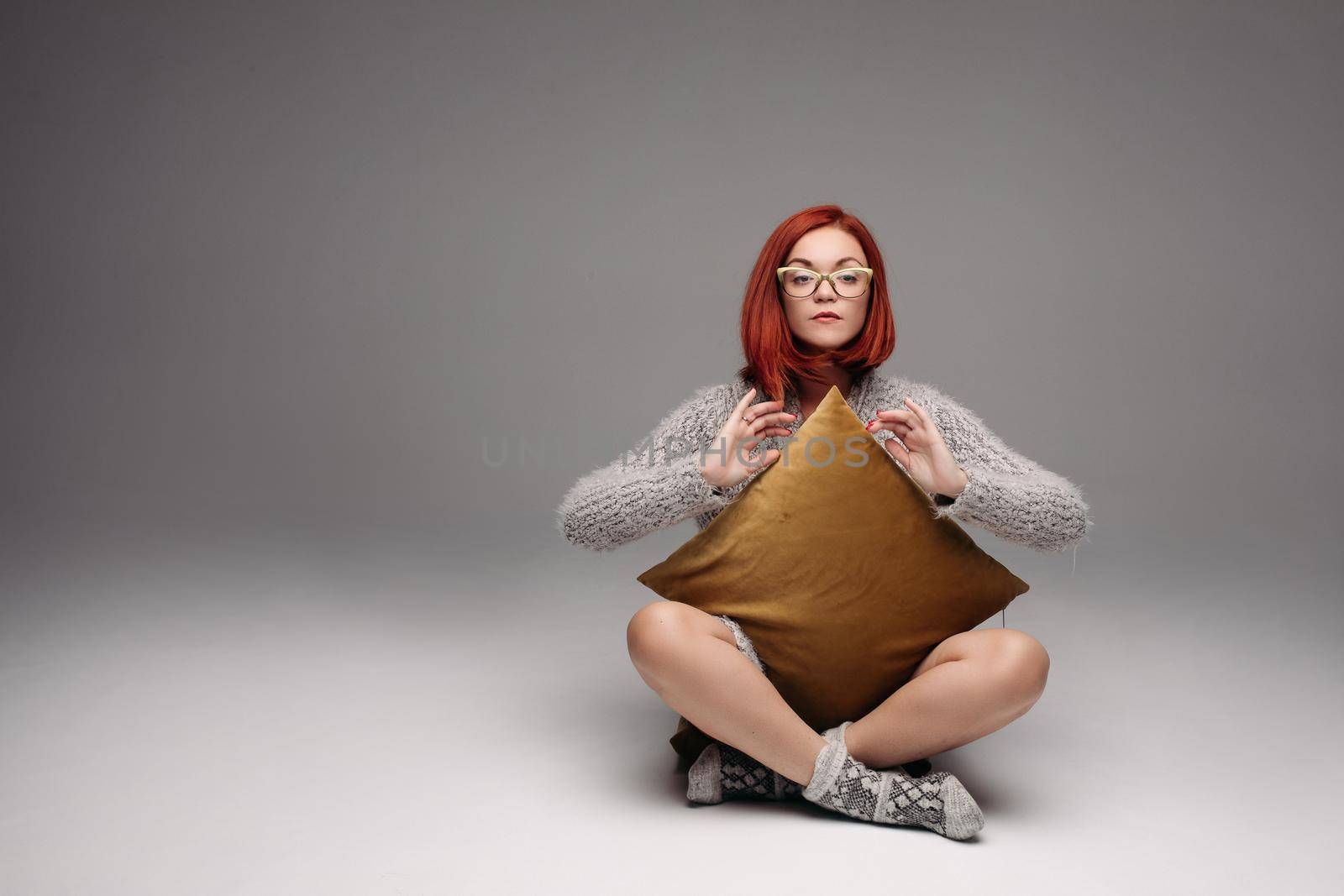Red haired girl in grey sweater and warm socks sitting on floor in Turkish style and sadly looking at camera. Beautiful girl in glasses with cross on her neck holding pillow between hands and legs.