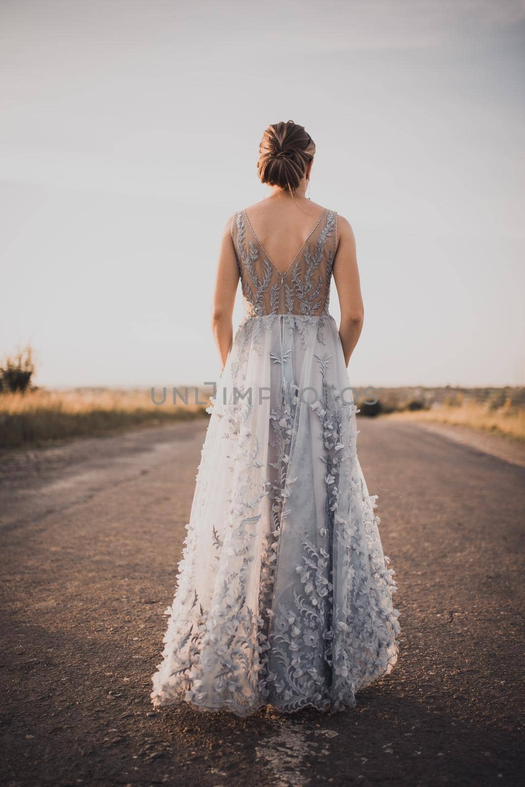 a young girl walks along an asphalt road in a gray blue long dress at sunset by AndriiDrachuk