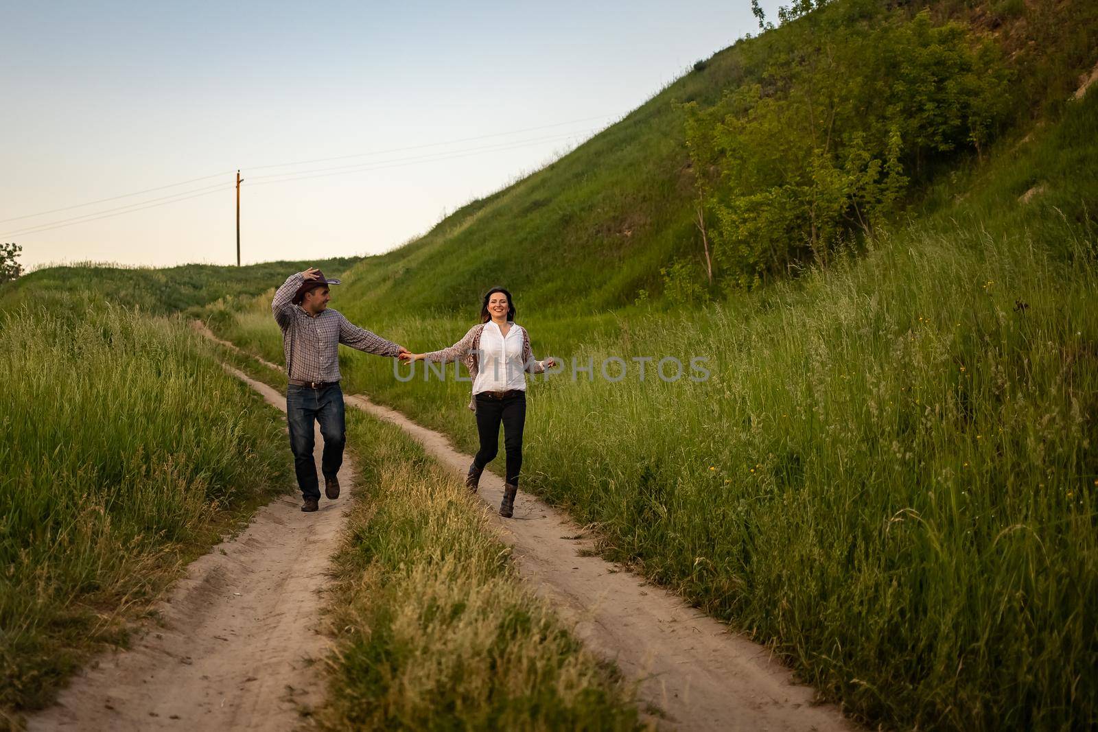 a couple in love runs holding hands along the road in the village. Guy in a cowboy hat by Anyatachka