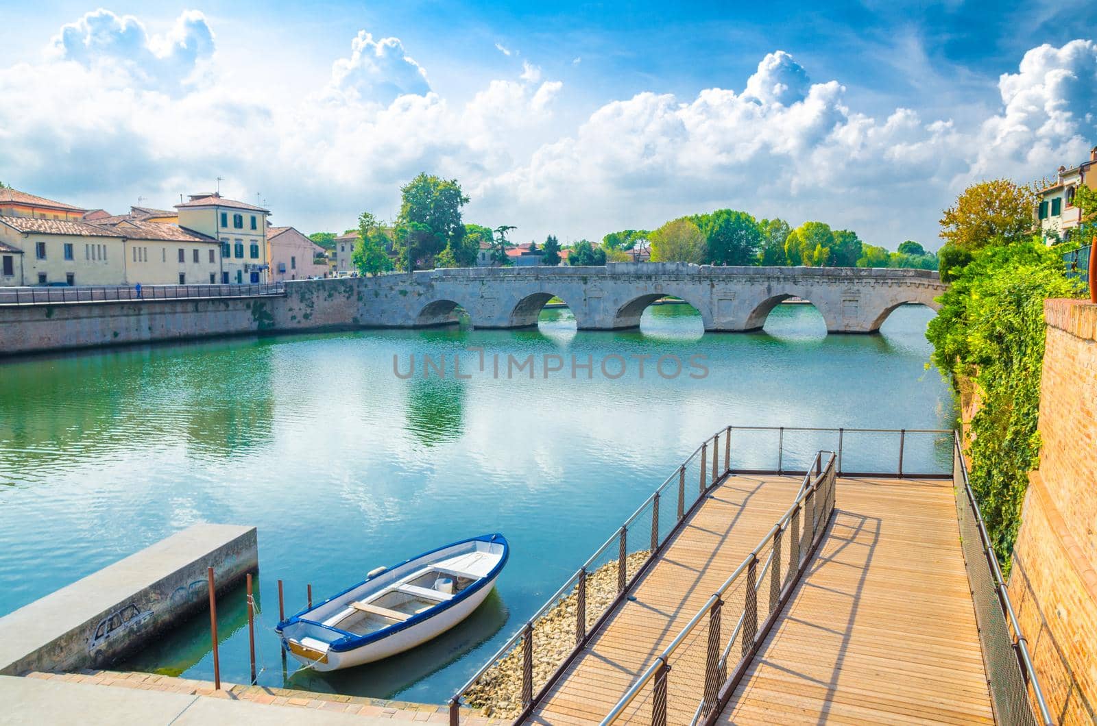 Boat in blue water near pier and stone arch Tiberius bridge Ponte di Tiberio, Augustus Bridge over Marecchia river in historical city centre Rimini with blue sky white clouds, Emilia-Romagna, Italy