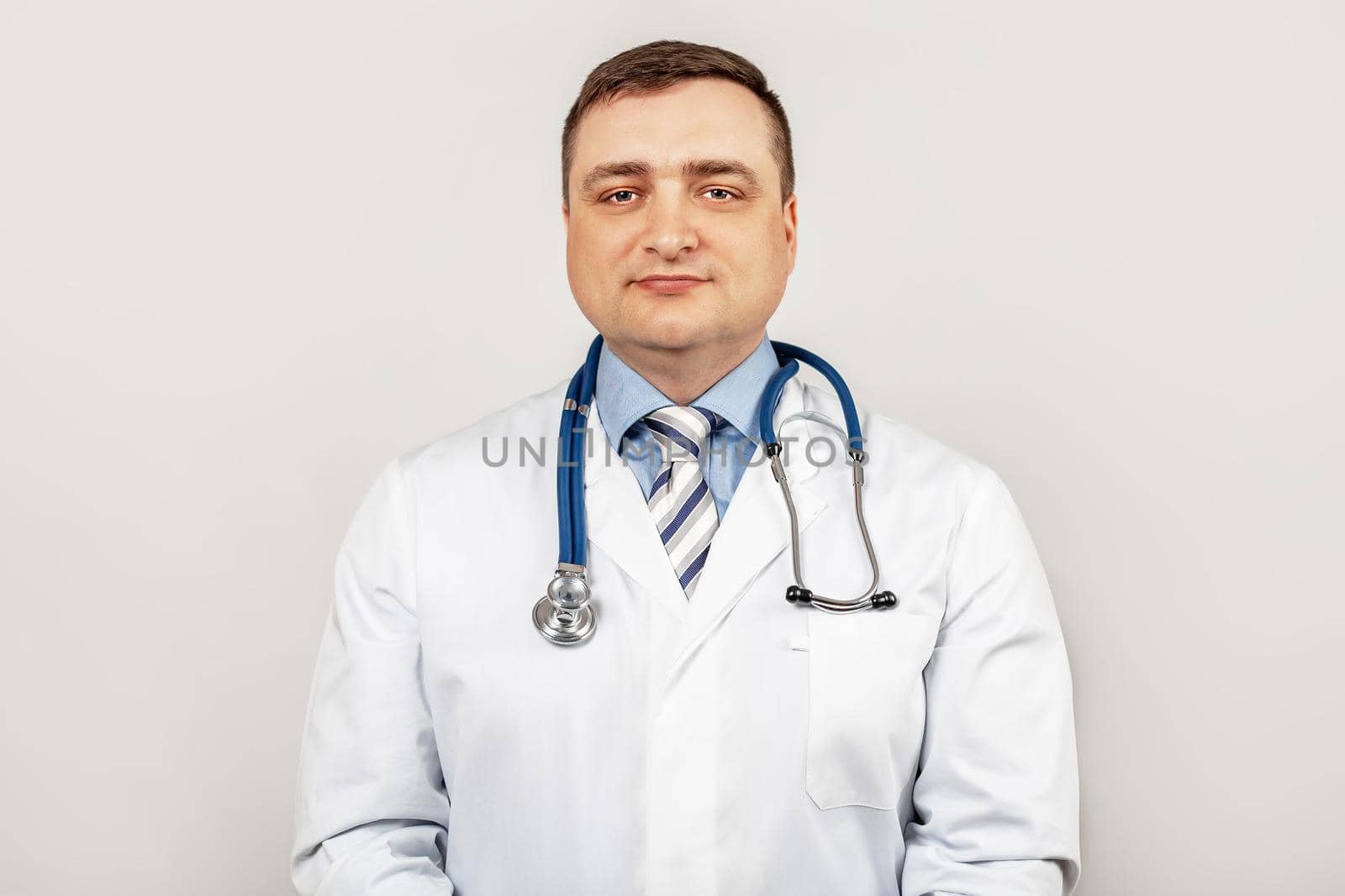 Portrait of smiling young male doctor with a stethoscope around neck standing within a white coat, isolated on gray background