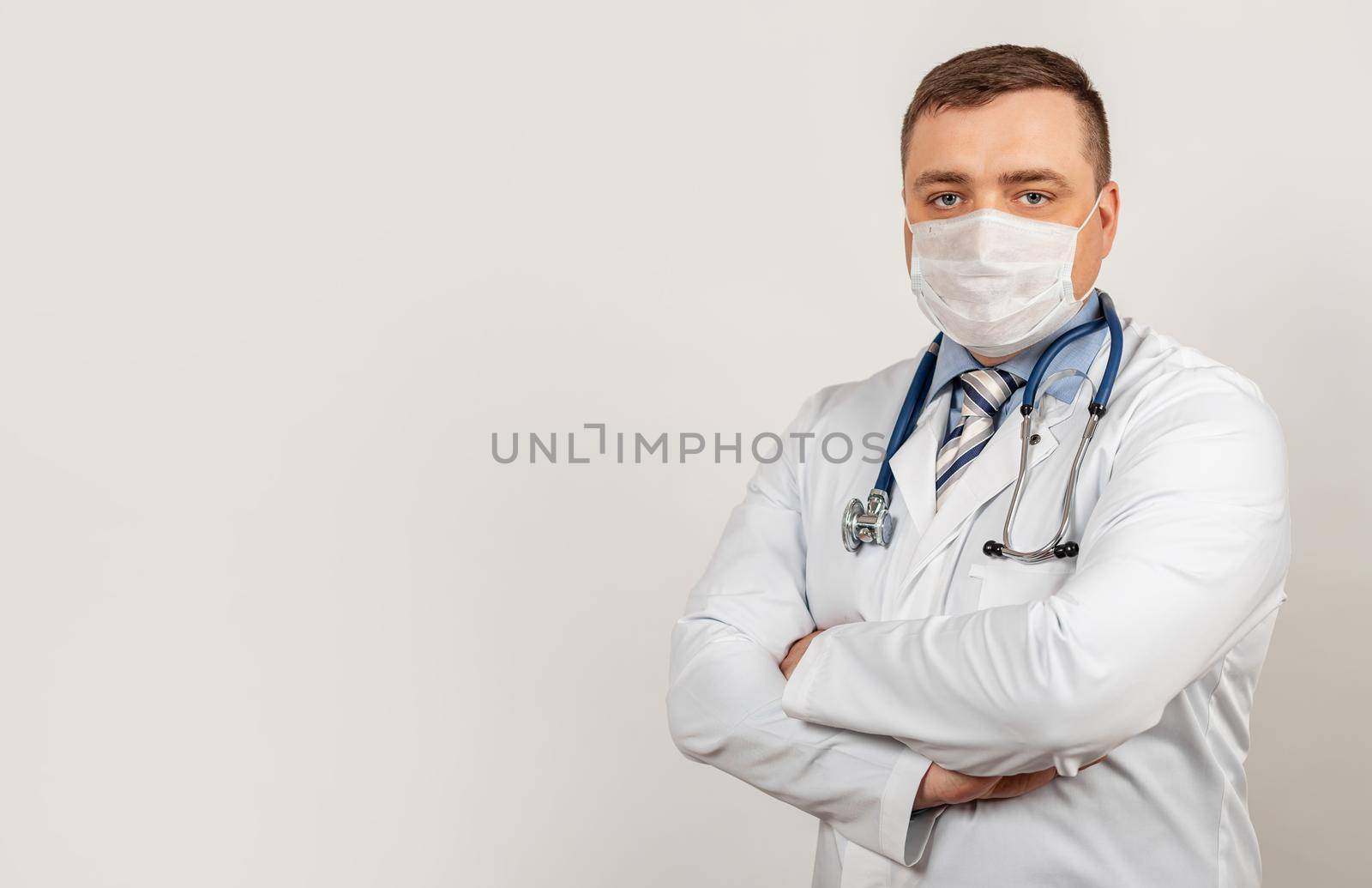 A male doctor in a medical mask with arms crossed, on his chest poses in front of the camera in a white coat near a white building during the COVID-19.