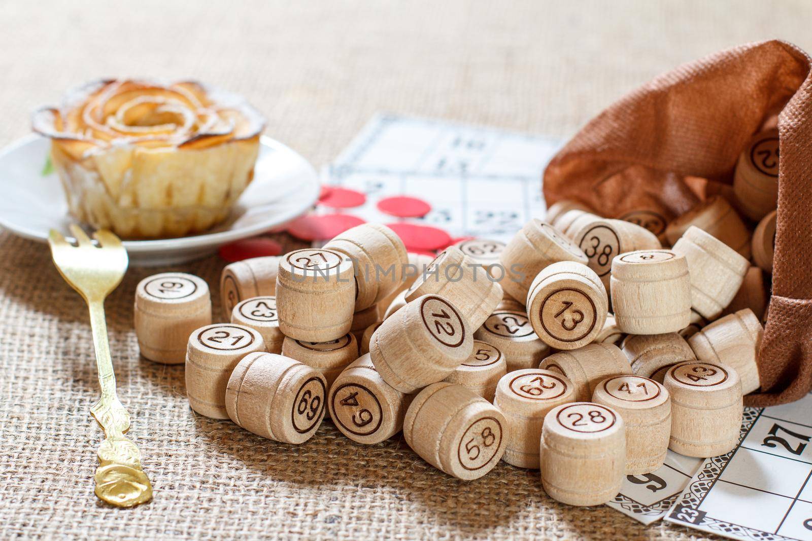 Wooden lotto barrels in brown pouch and game cards for a game in lotto with homemade biscuit in the form of rose on white saucer with fork on the background. Selective focus on barrels