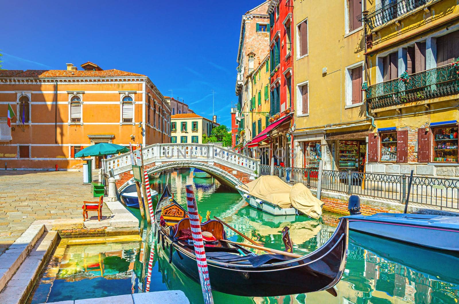 Venice cityscape with gondola and motor boats moored on narrow water canal Rio dei Frari, colorful buildings and stone bridge, Veneto Region, Northern Italy, blue sky background in summer day