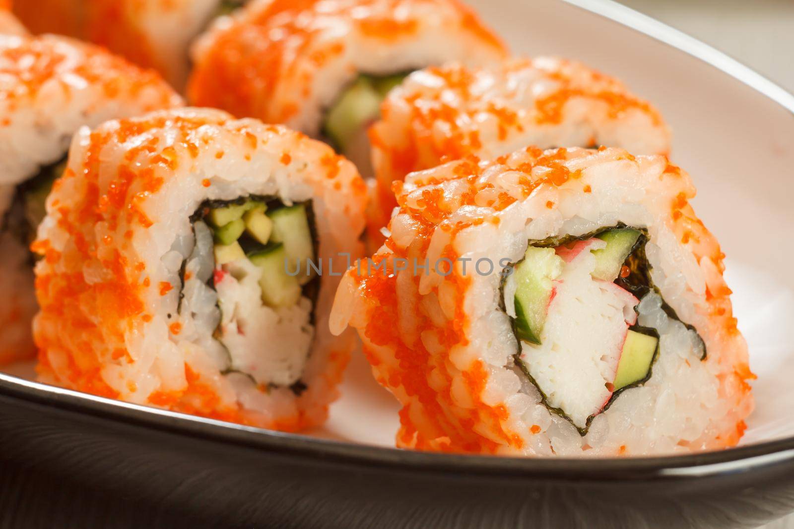 Close up Uramaki California. Sushi rolls with nori, rice, pieces of avocado, cucumber, crab sticks decorated with flying fish roe on ceramic plate. Japanese cuisine. Shallow depth of field