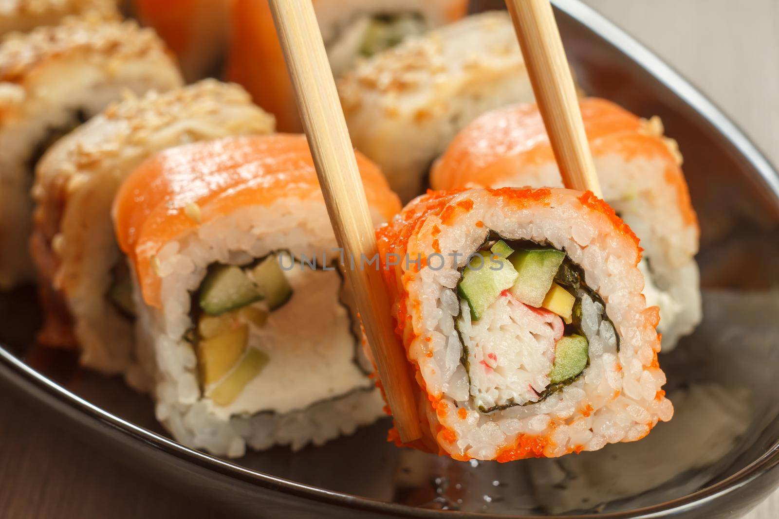 Two chopsticks holding Uramaki California and different sushi rolls with seafood on ceramic plate on the background. Japanese cuisine. Shallow depth of field. Focus on roll with chopsticks