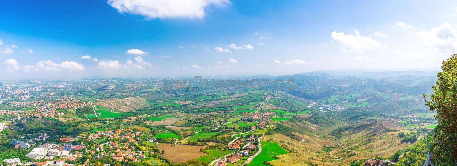 Aerial top panoramic view of landscape with valley, green hills, fields and villages of Republic San Marino suburban district with blue sky white clouds background. View from San Marino fortress.