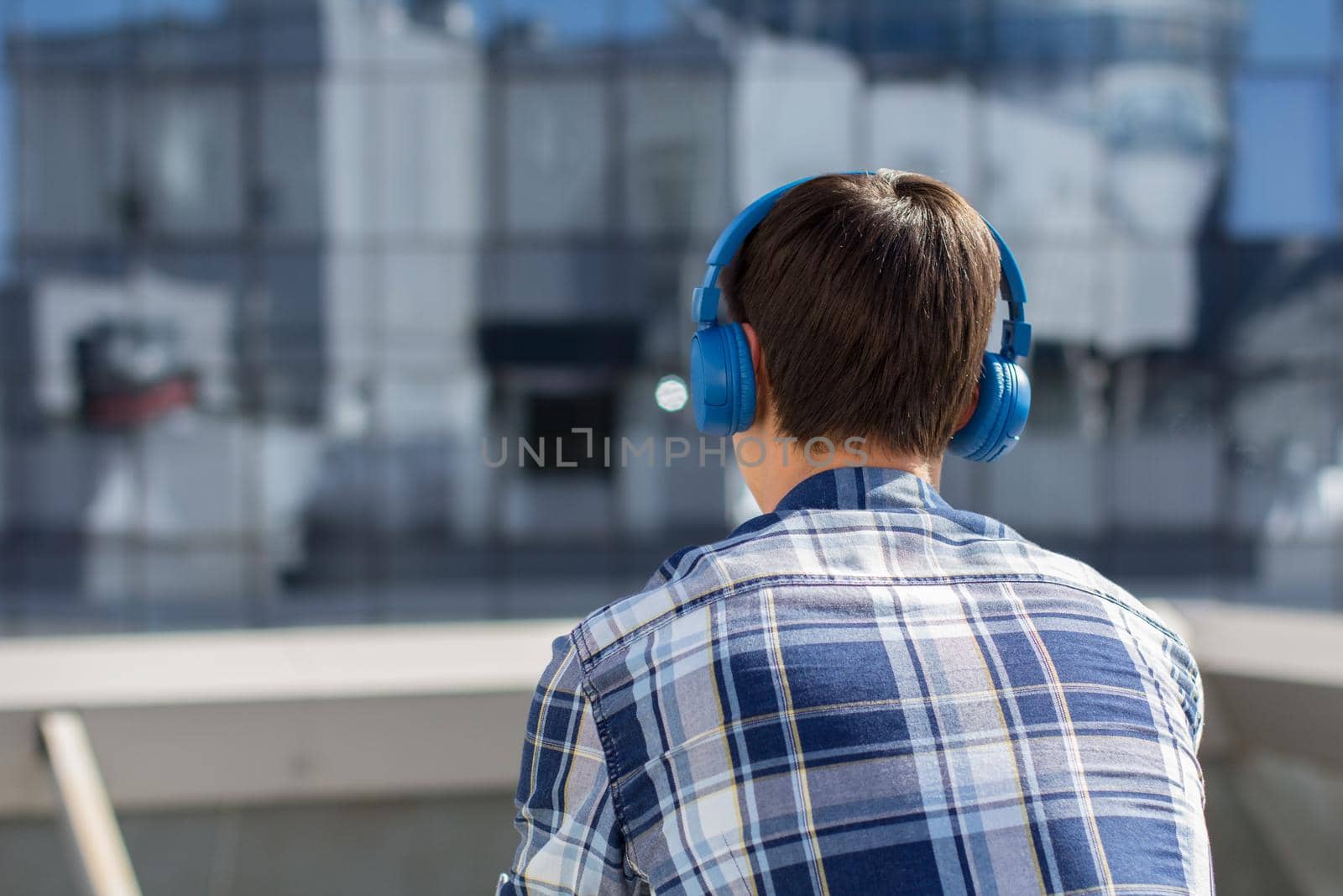 Back view of young man in checkered shirt with wireless headphones looking at cityscape. Guy listening to music on headphones in front of modern building