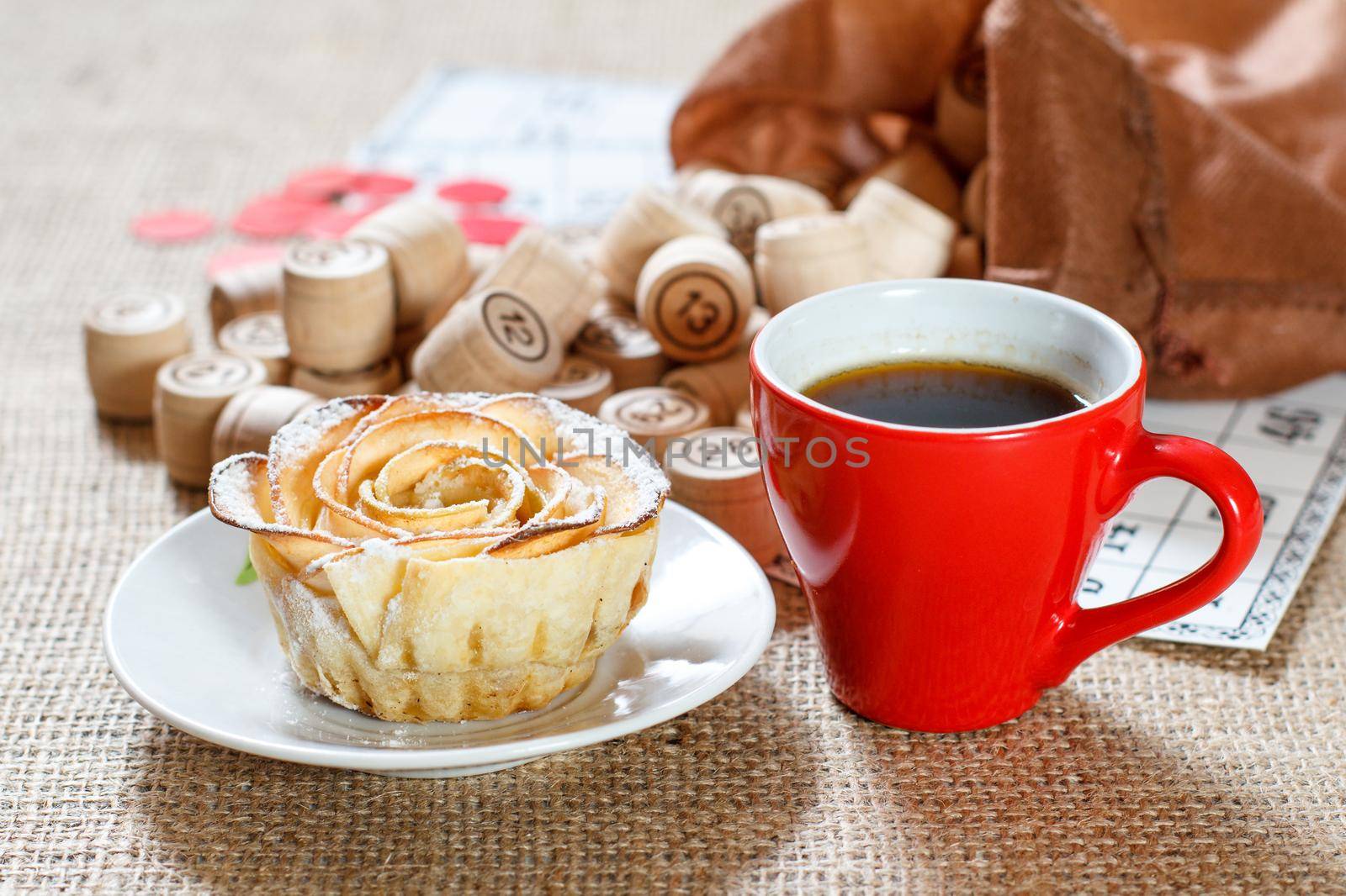Wooden lotto barrels in brown pouch and game cards for a game in lotto with cup of coffee and homemade biscuit in the form of rose on white saucer
