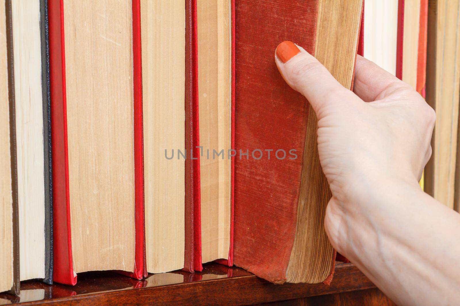 Female hand is taking an old book from the bookshelf. Many hardback books on wooden shelf. Library concept