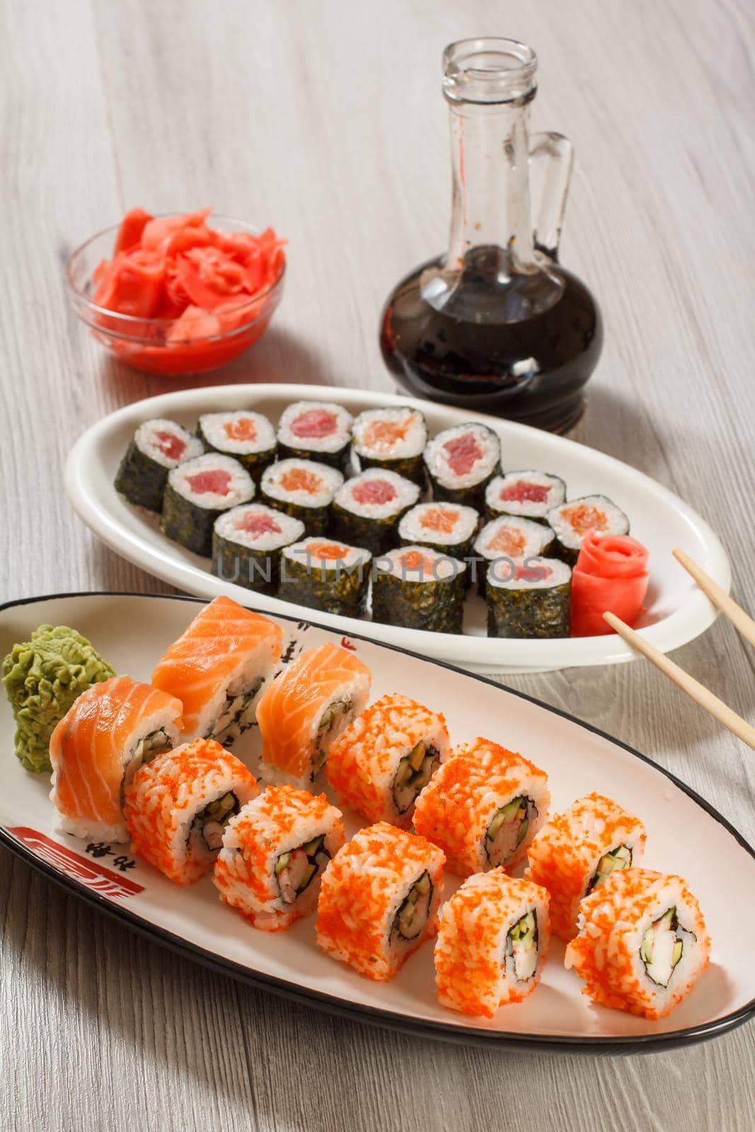 Different sushi rolls with rice, vegetable, seafood on ceramic plates, glass bottle with soy sauce and red pickled ginger in a bowl on wooden desk