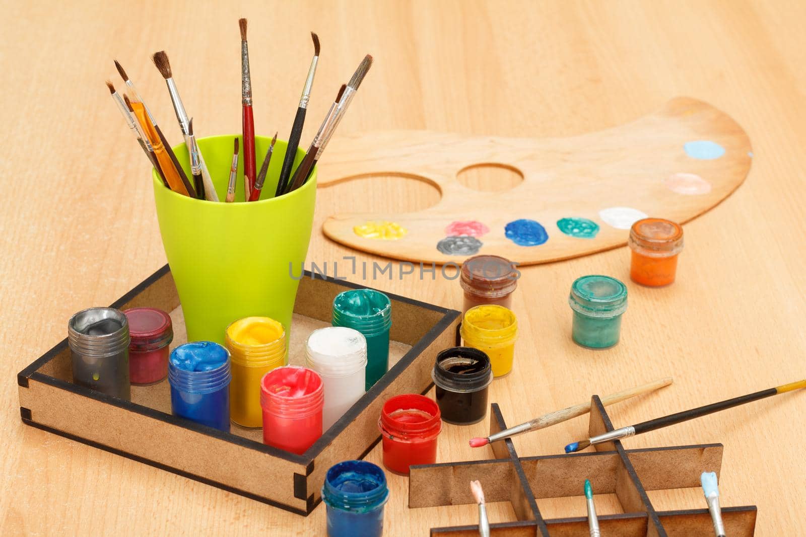 Wooden palette with paints of different colors and cup with paintbrushes in box on the table