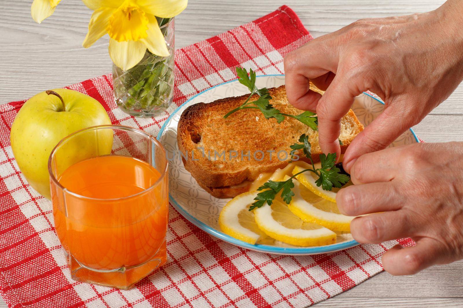 Female hands lay down breakfast food on a plate. Toast with butter and cheese, slices of lemon on plate, apple, bouquet of yellow daffodils and glass of orange juice on kitchen napkin