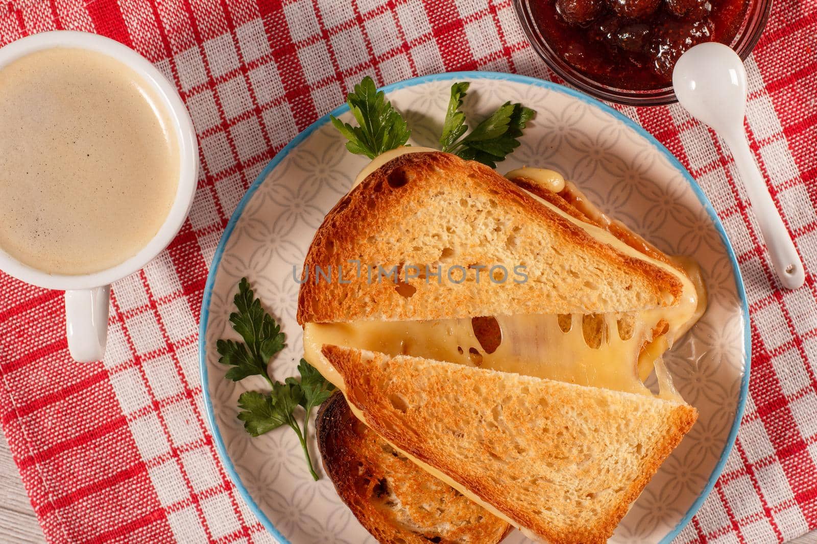 Toasted slices of bread with cheese and green parsley on white plate, cup of coffee, glass bowl with strawberry jam and spoon with red kitchen napkin. Top view. Good food for breakfast