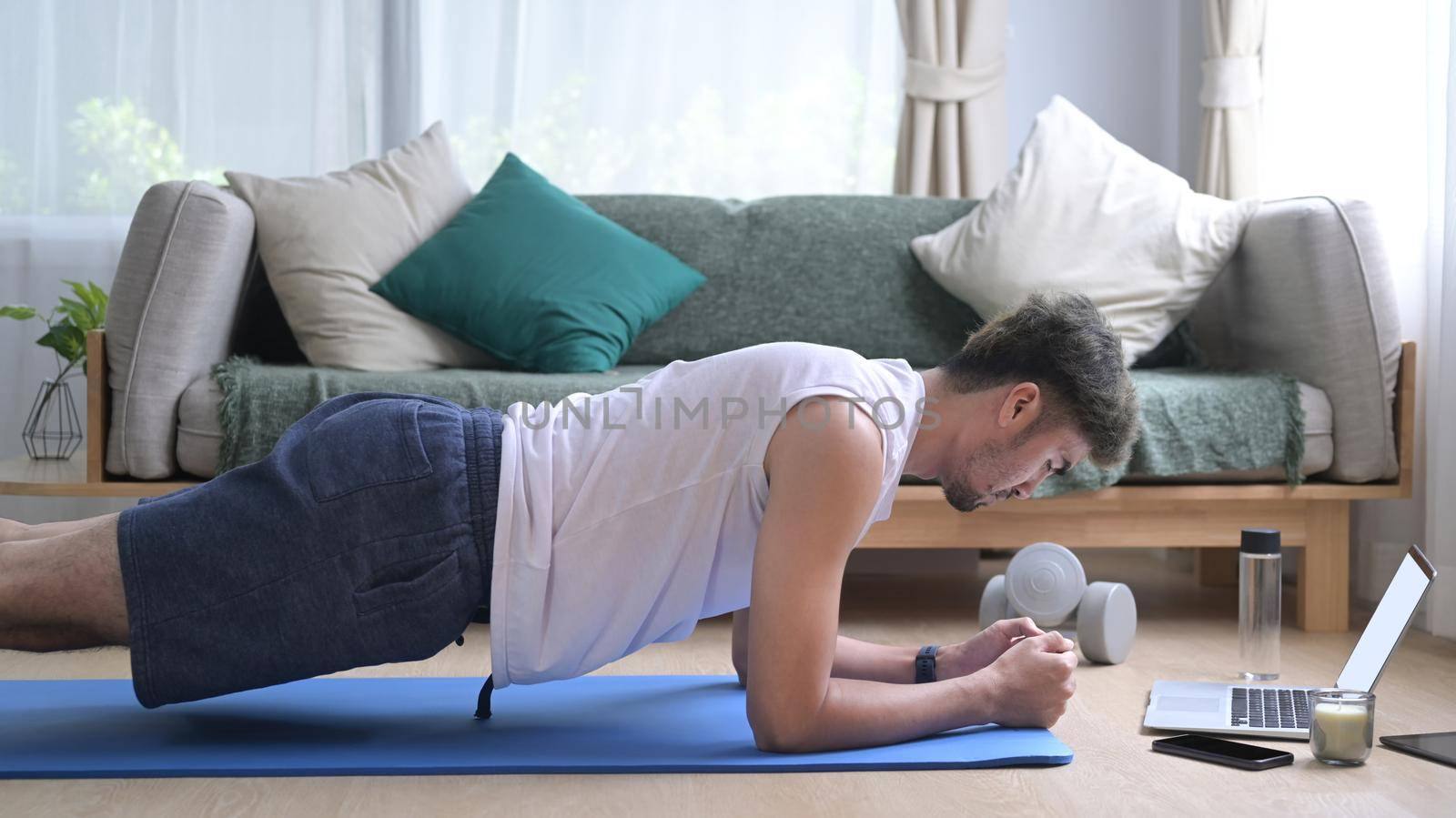 Young man in sports clothes doing plank on blue mat and watching fitness lessons online on laptop. Sport at home, workout, fitness and exercises.