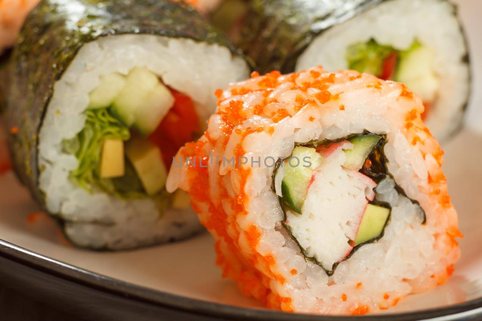 Close up Uramaki California. Sushi roll with nori, rice, pieces of avocado, cucumber, crab sticks decorated with flying fish roe and sushi rolls with vegetables on the background. Japanese cuisine. Shallow depth of field