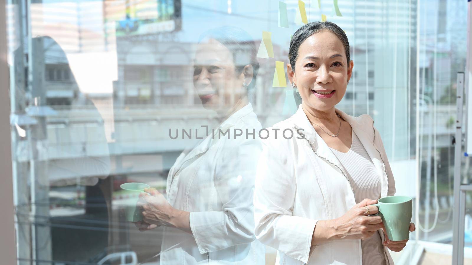 Confident middle aged business leader standing near large window at office with city buildings in background.