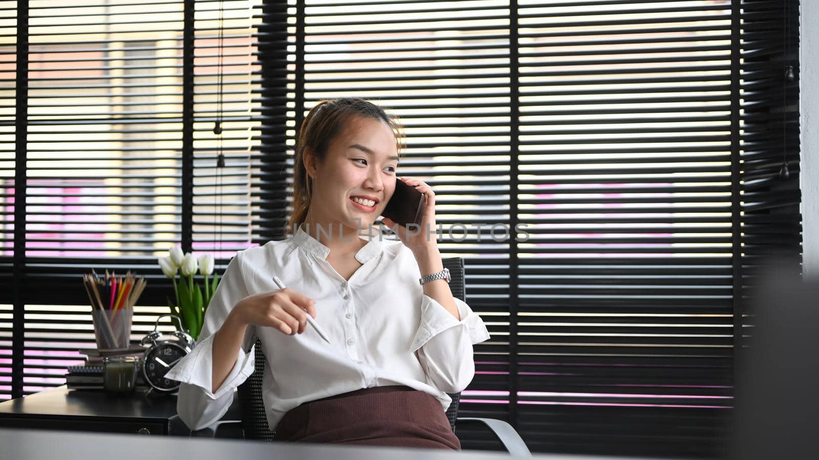 Satisfied young businesswoman sitting at her workplace and talking on cellphone.