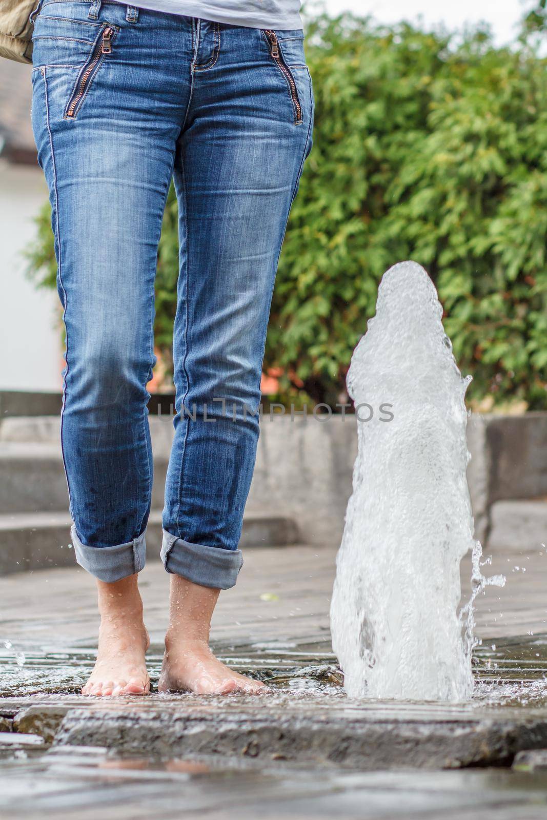 Feet of the lady in jeans with drops of water on them near the fountain in city. Sunny day in summertime