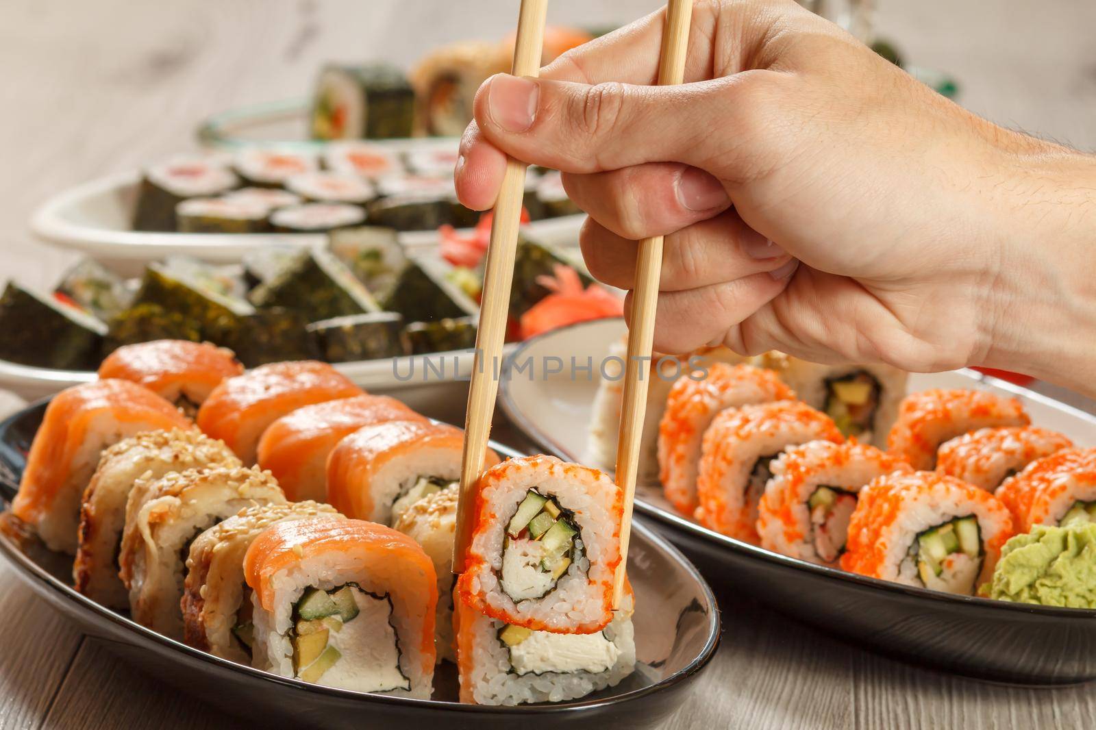Male hand with two chopsticks holding Uramaki California roll and different sushi rolls with seafood on ceramic plates on the background. Japanese cuisine. Focus on roll with chopsticks