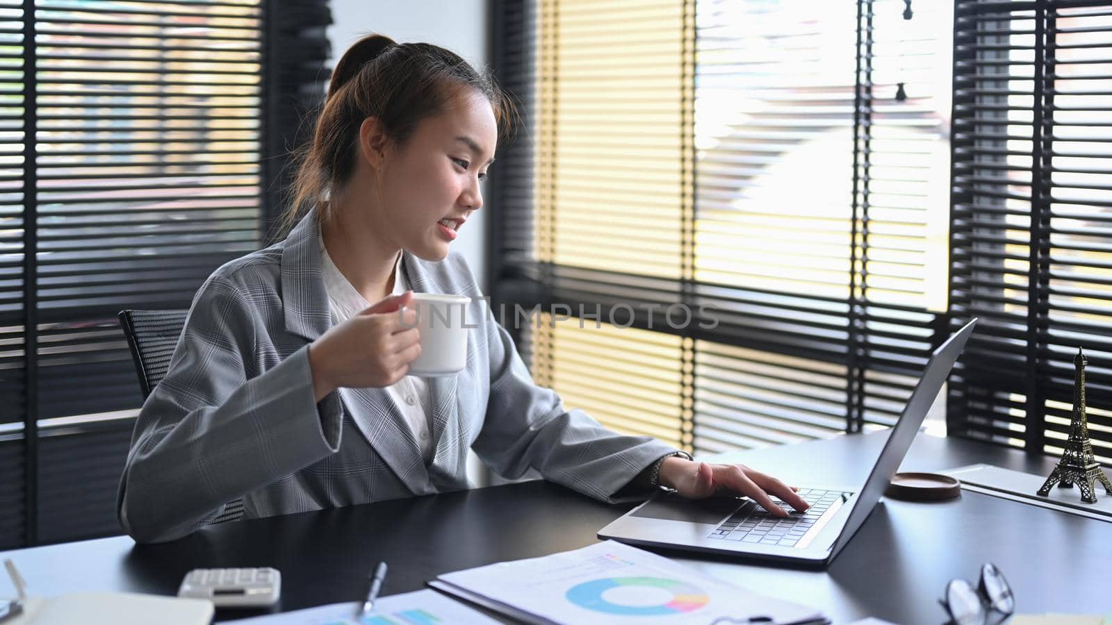 Professional chartered accountant drinking coffee and working with laptop computer at office desk.