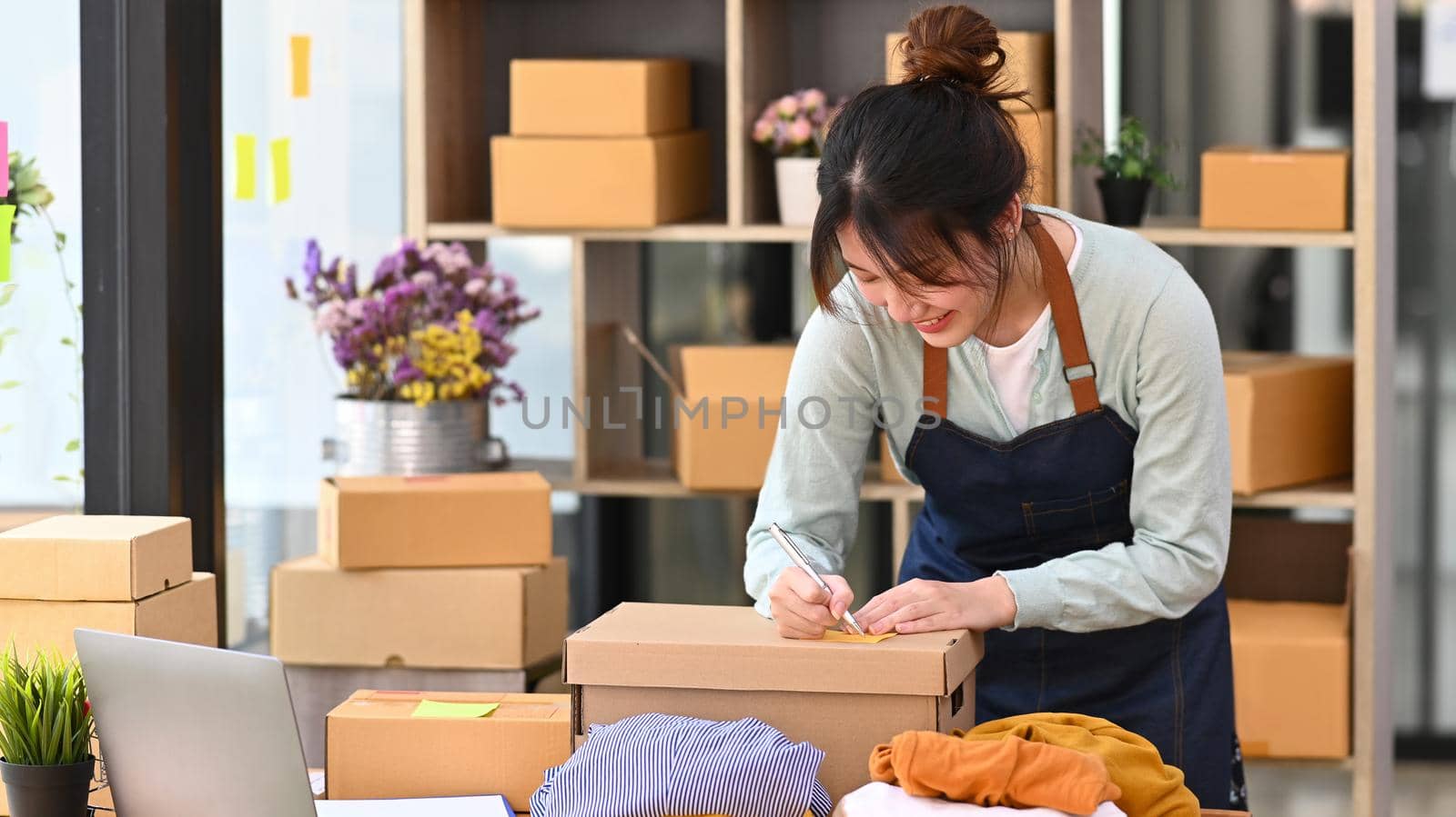 Happy young woman online seller writing address on cardboard box while preparing boxes of product for delivery. Online selling, E-commerce, Online Shopping.