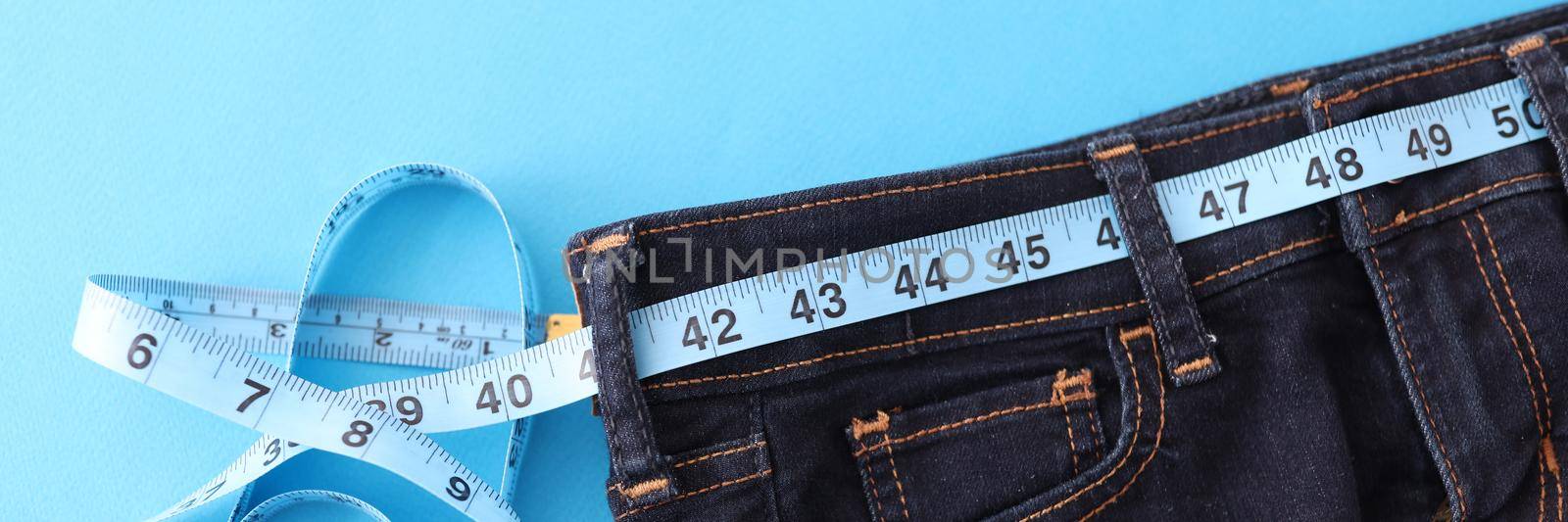 Measuring sewing tape lies on jeans on a blue background, close-up. Measurement of clothing size, tailoring and repair in the atelier