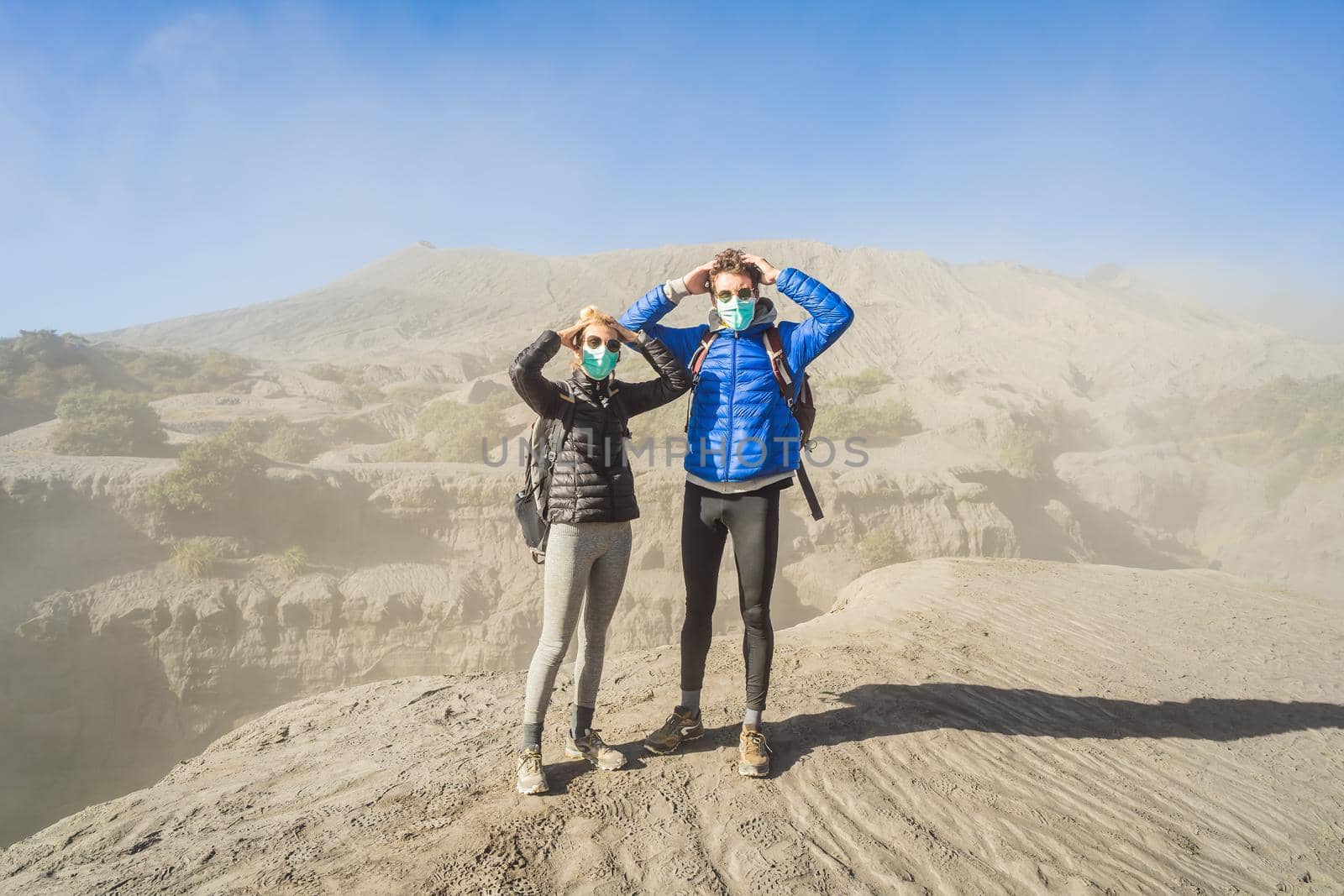 Young couple man and woman visit the Bromo volcano at the Tengger Semeru National Park on Java Island, Indonesia. They are scared of the eruption covering everything with dust and they wear safety masks to protect themselves from dangerous volcanic ash. Concept of tourists in trouble caused by a volcano.