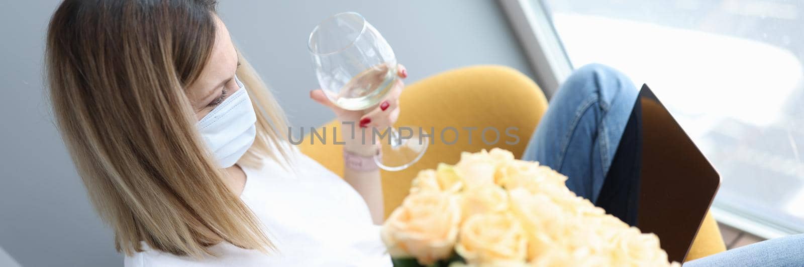 A woman in a mask sits by the window with a glass of wine and flowers. Celebration during lockdown, virus isolation