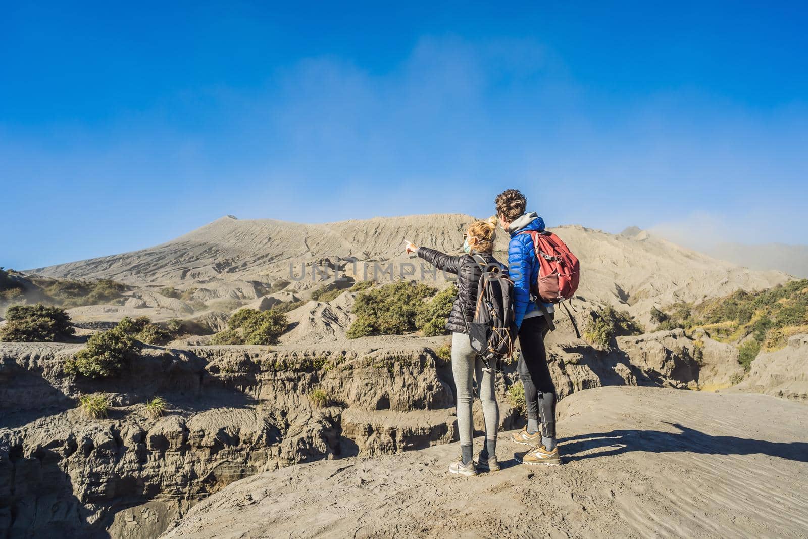 Young couple man and woman visit the Bromo volcano at the Tengger Semeru National Park on Java Island, Indonesia. They enjoy the magnificent view on the Bromo or Gunung Bromo on Indonesian, Semeru and other volcanoes located inside of the Sea of Sand within the Tengger Caldera. They wear safety masks to protect themselves from dangerous volcanic ash.